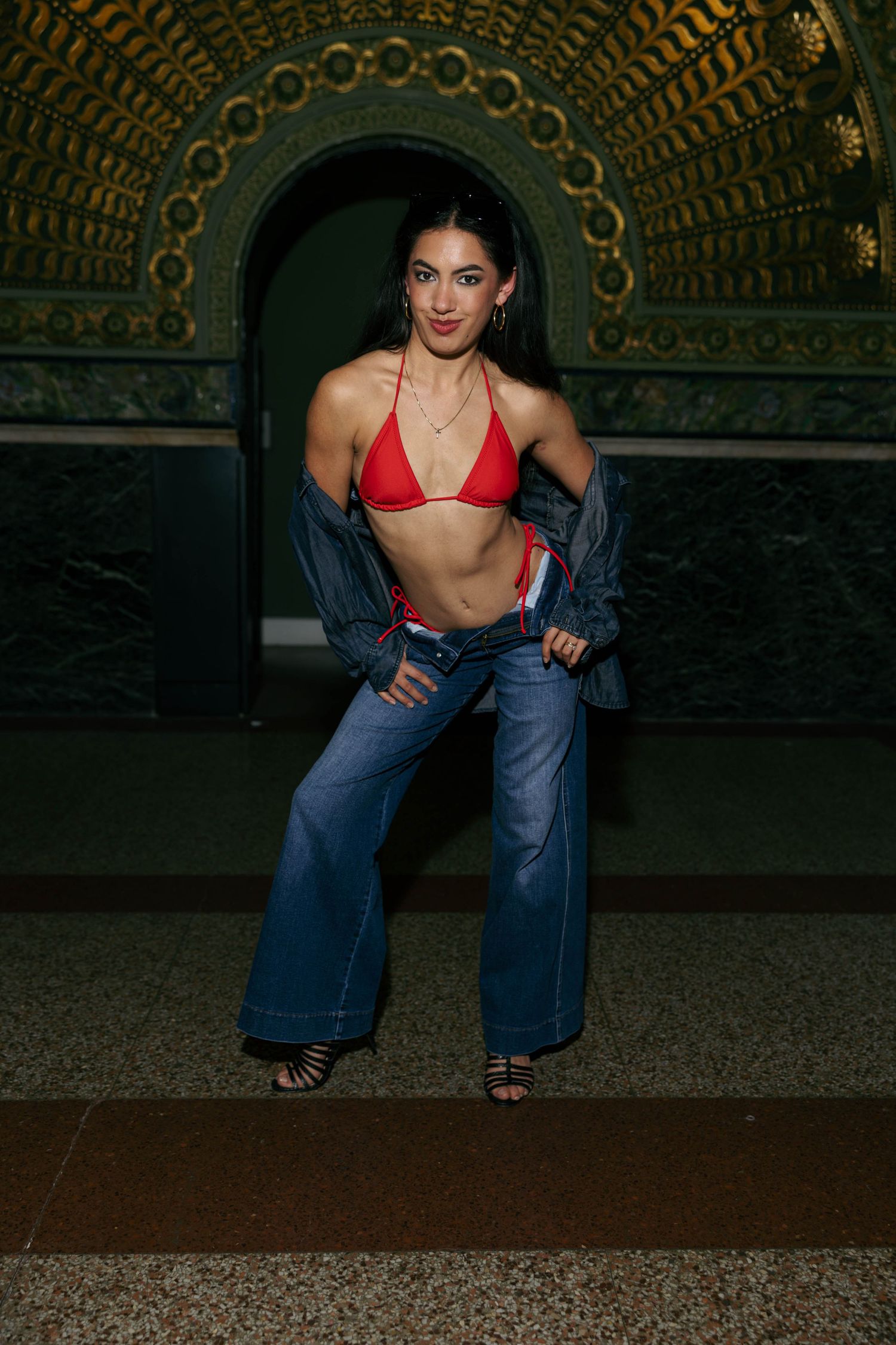 Woman in red bikini top, jeans, and jacket posing in ornate green archway.