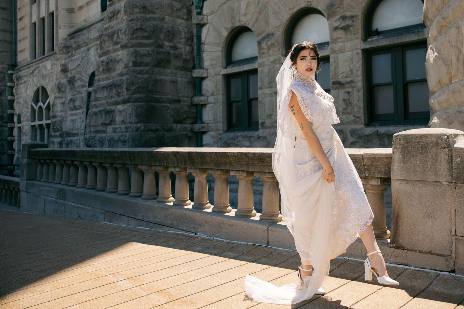 Elegant bride in a white dress and veil posing on a sunlit stone balcony of a historic building.
