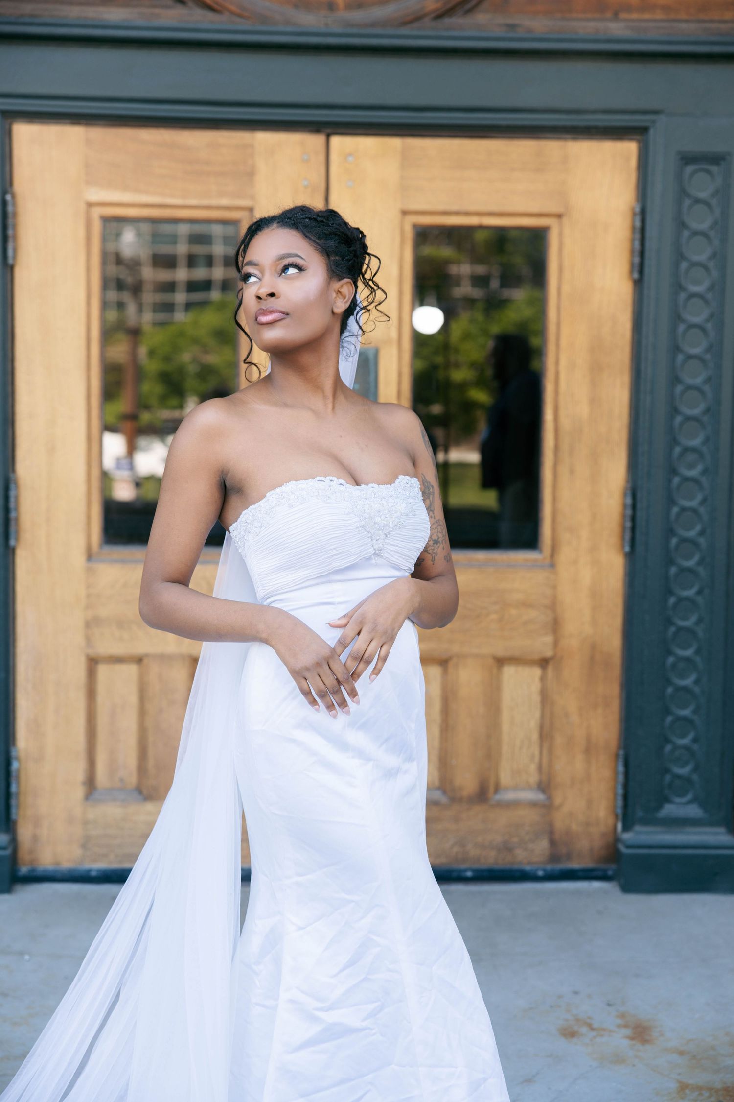 Elegant Black bride in strapless white mermaid gown with cape poses before warm wooden church doors.