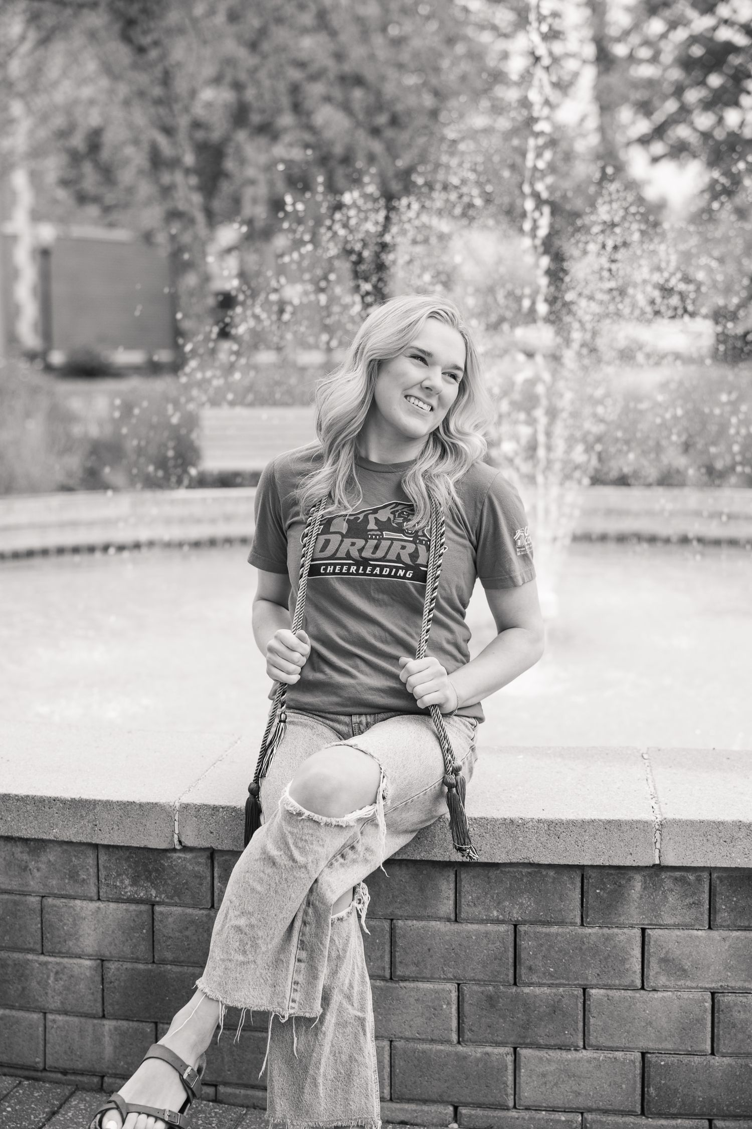 Person in university t-shirt sitting by fountain with water spraying in background.