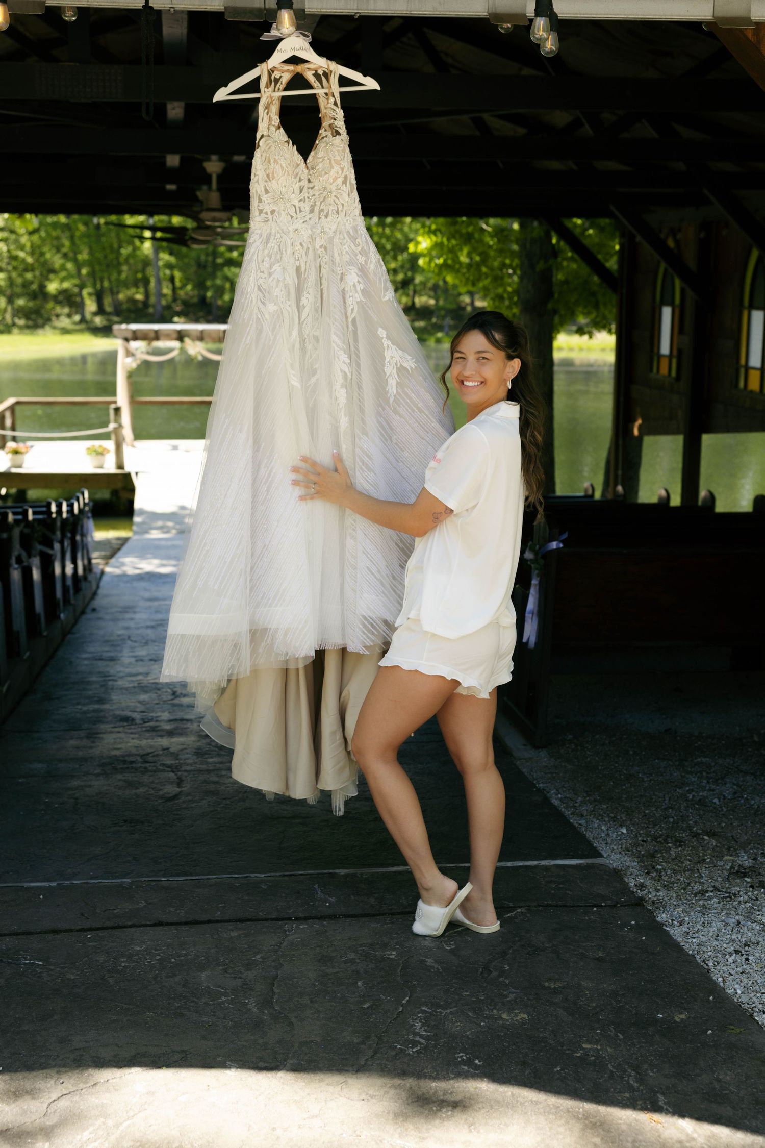 Smiling bride in white outfit holding her lace wedding gown on a hanger at an outdoor venue near a lake.