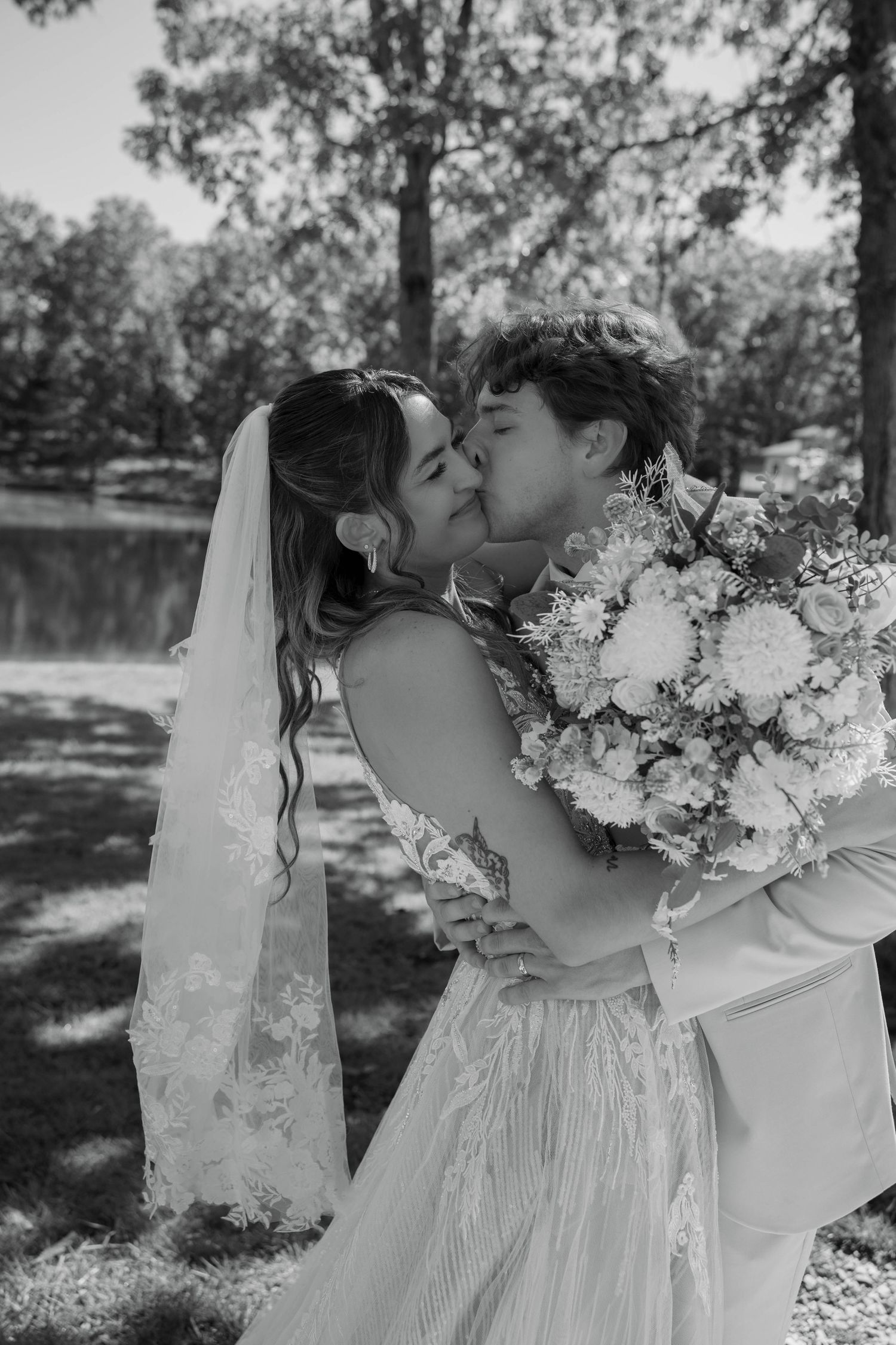 Black and white photo of a bride and groom sharing a kiss outdoors, bride holding a bouquet near a serene pond.