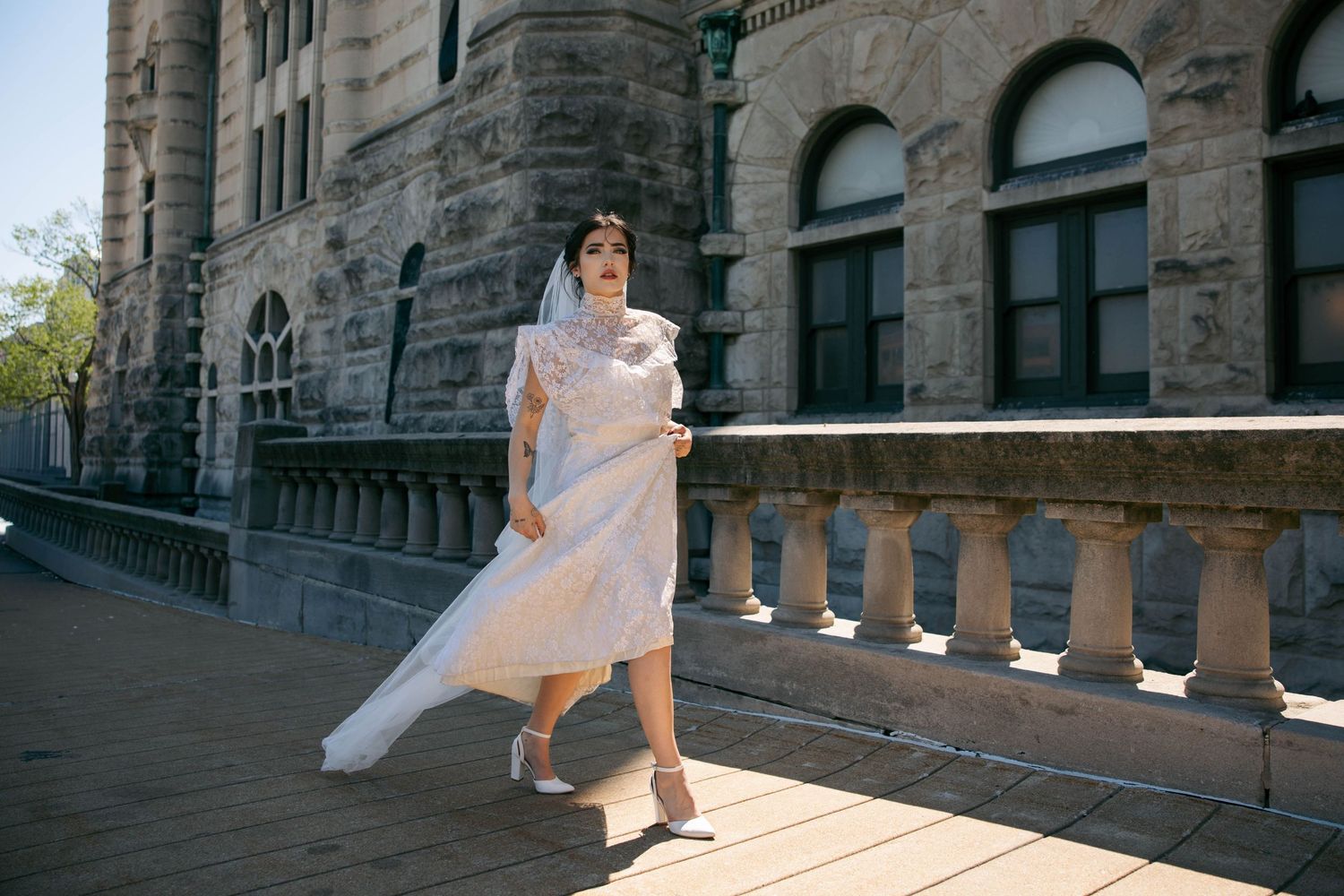 Elegant bride in short lace dress and veil walking on sunny stone balcony.