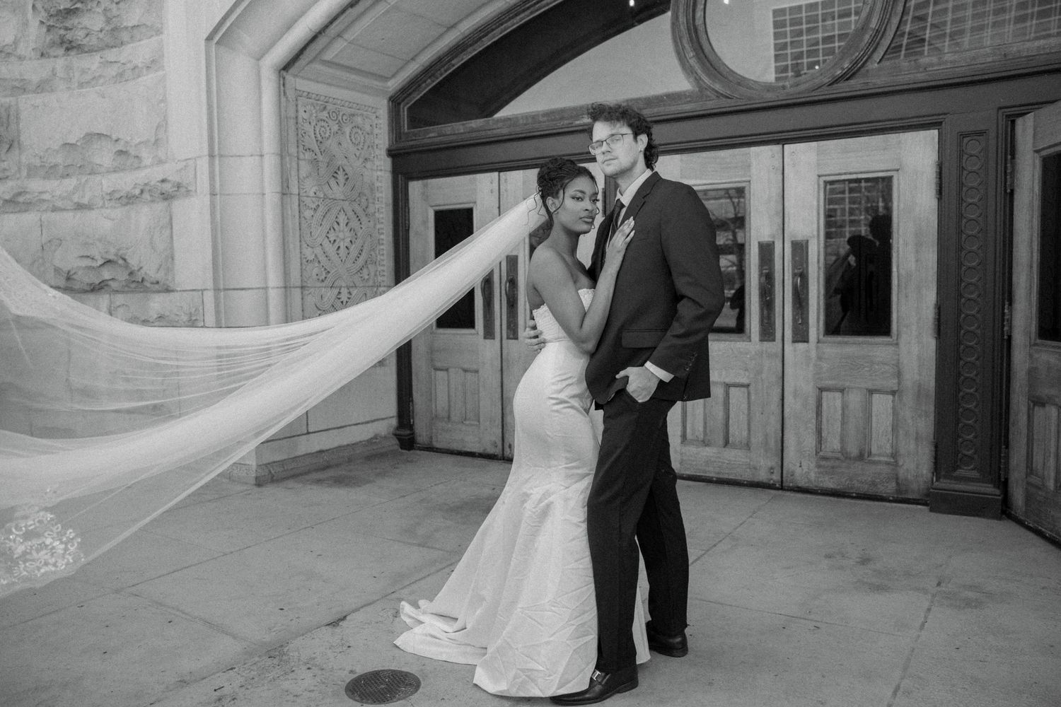 Bride and groom embrace outside ornate building entrance with long flowing cathedral veil, black and white photo.