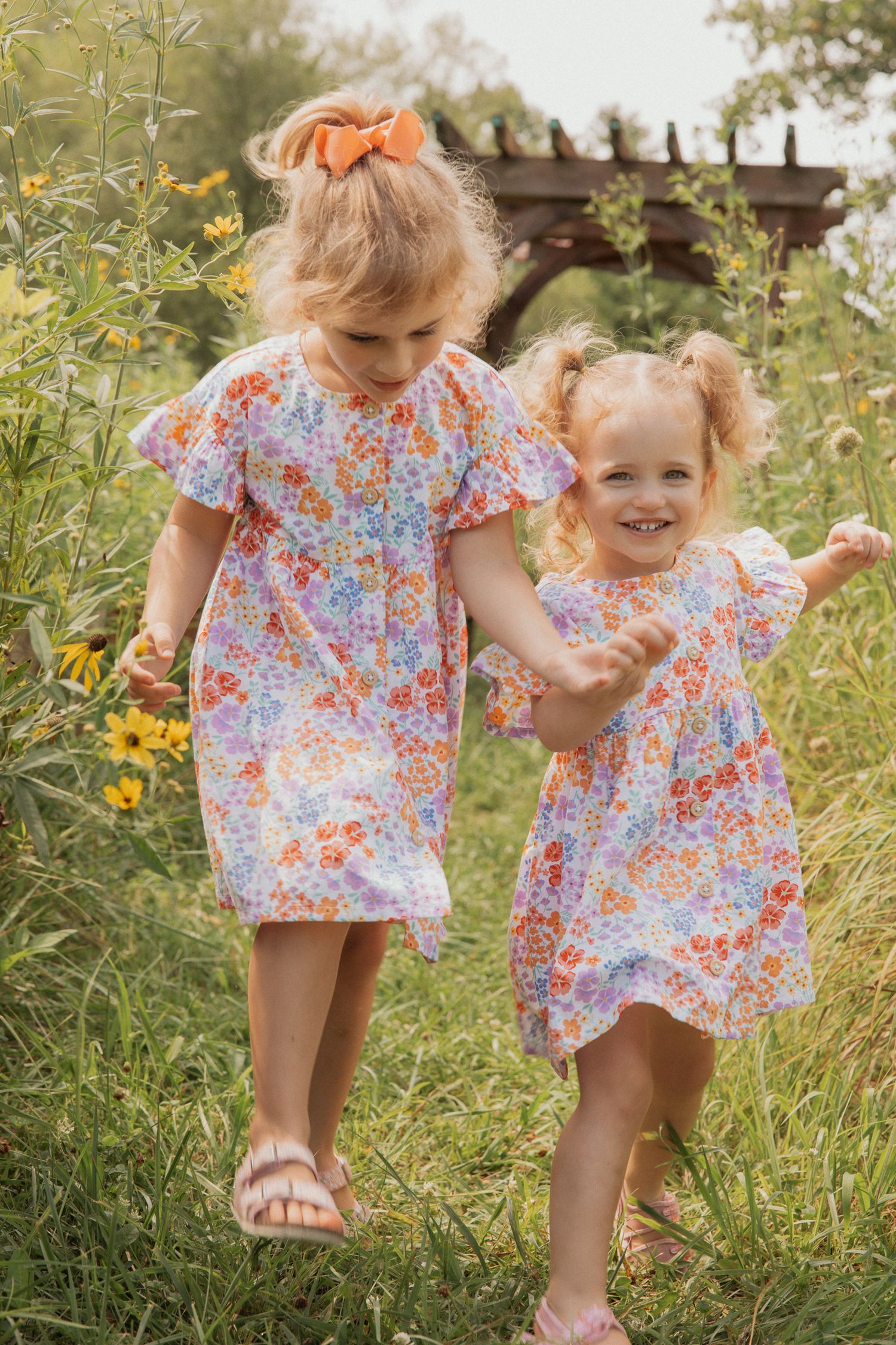 Two children in matching floral dresses walking together through summer wildflowers.