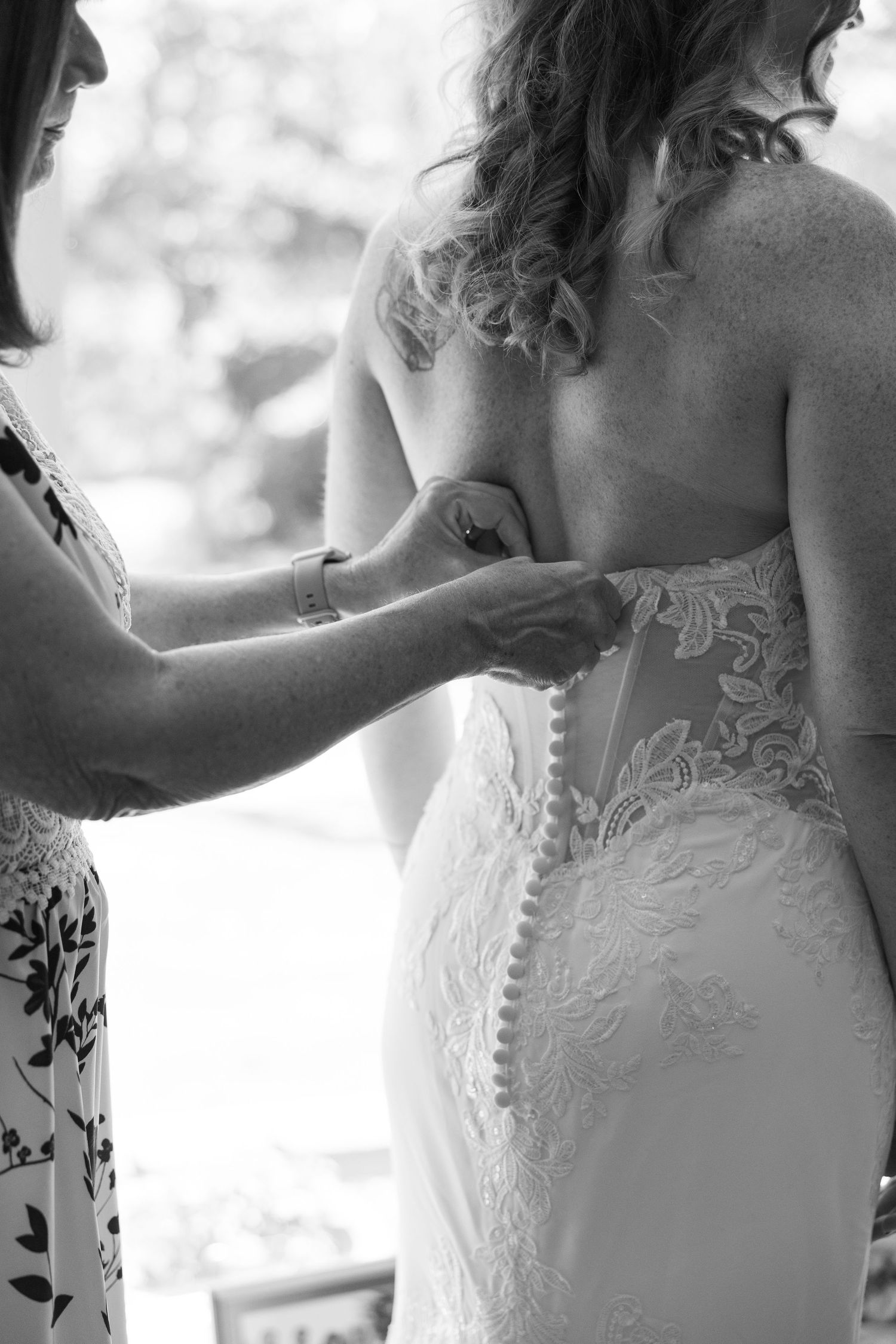 Black and white image of wedding dress being buttoned up showing intricate lace details.