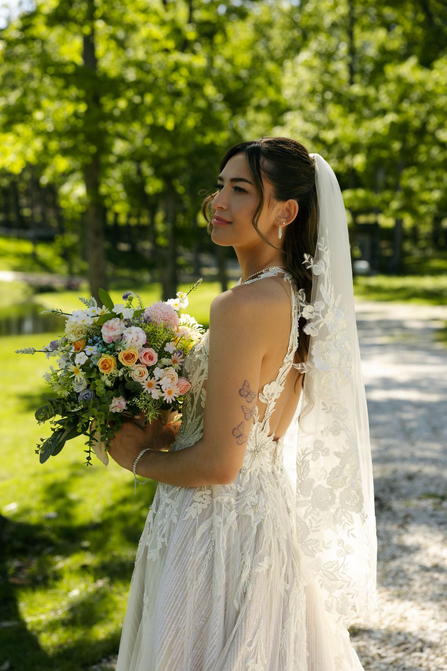 Bride in lace backless wedding gown holding colorful bouquet, standing in sunlit garden with veil.