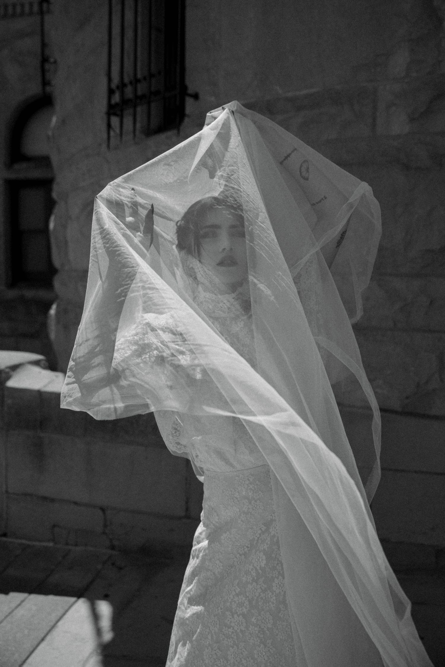 Bride in lace wedding dress with flowing veil caught by wind, black and white photo outside stone building.