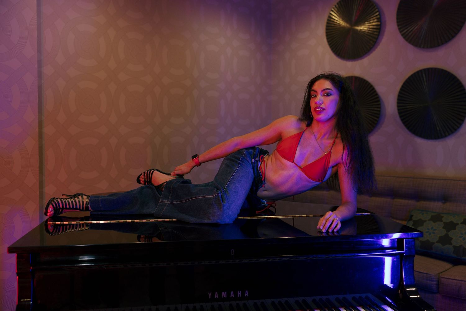 Woman in red bralette and jeans reclining on grand piano in purple-lit room.