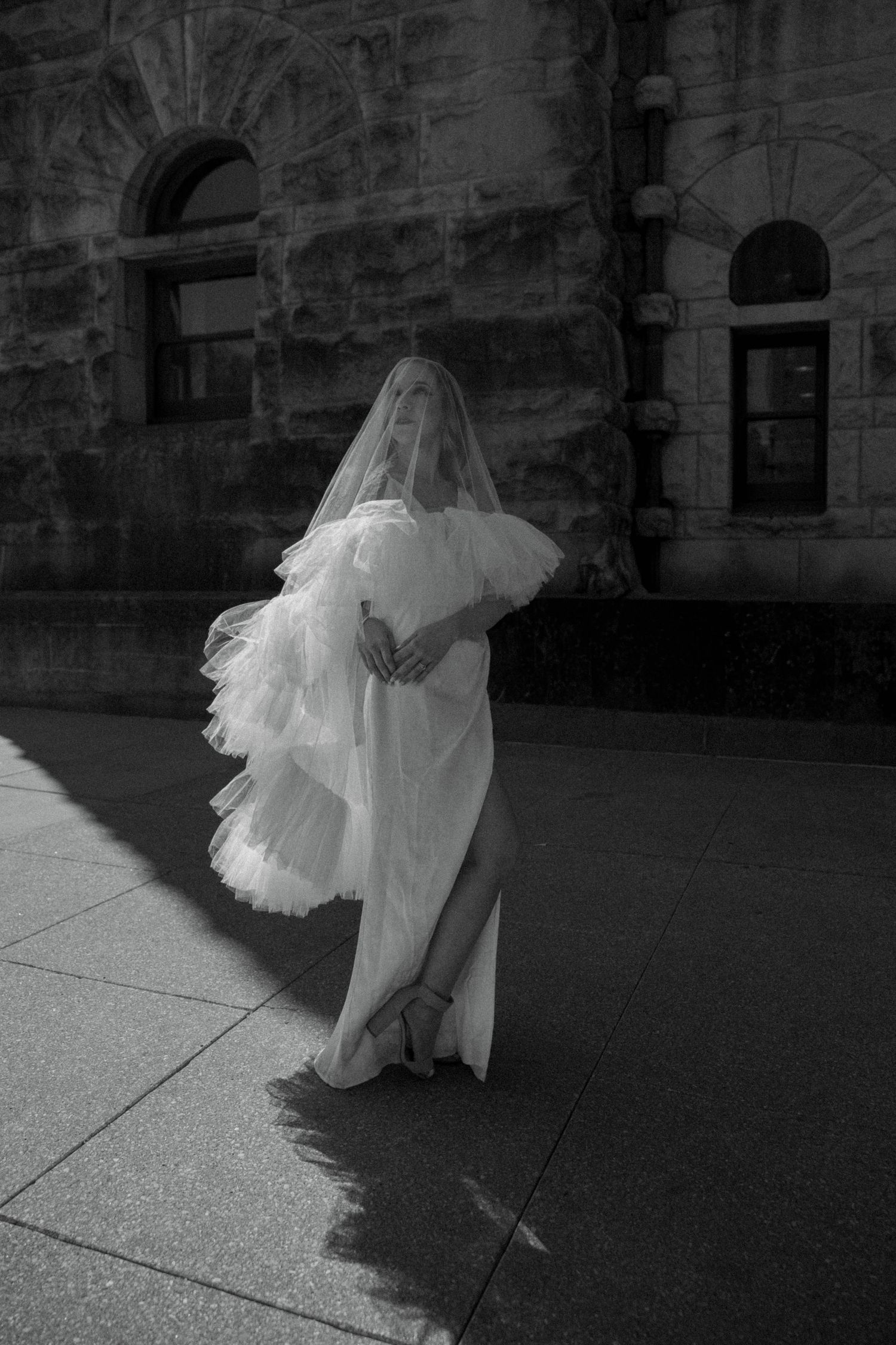 Bride in flowing white tulle gown and veil poses on sunlit sidewalk before a historic stone building.