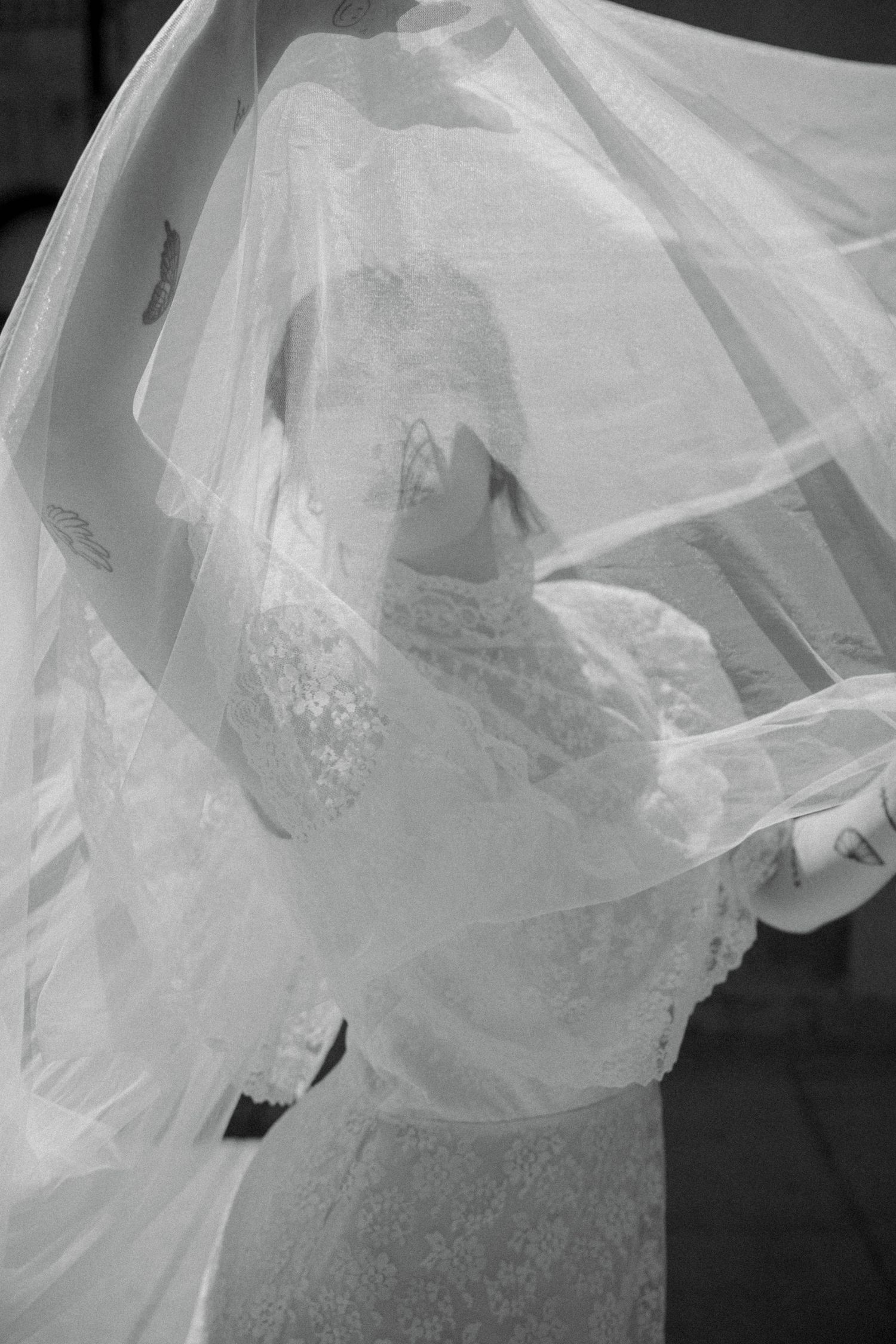 Bride in lace wedding dress with flowing veil caught in wind, elegant black and white bridal portrait.