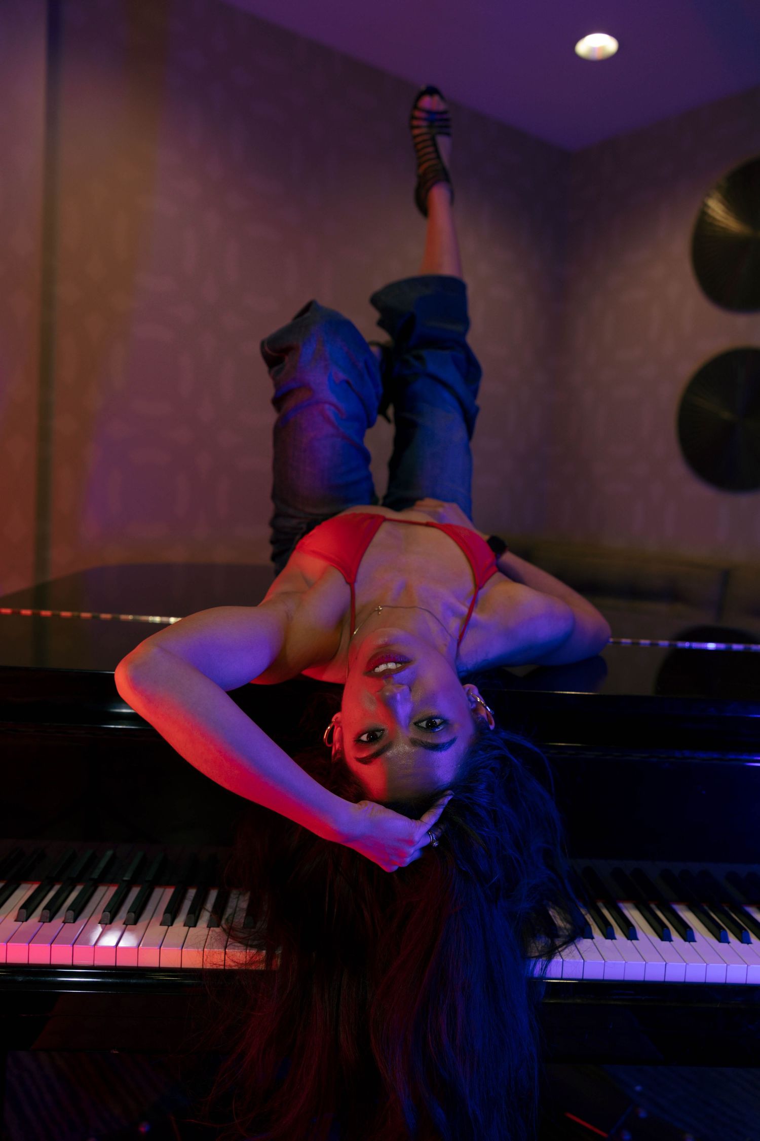 Woman in red top and jeans lying across a grand piano in moody purple and red studio lighting.