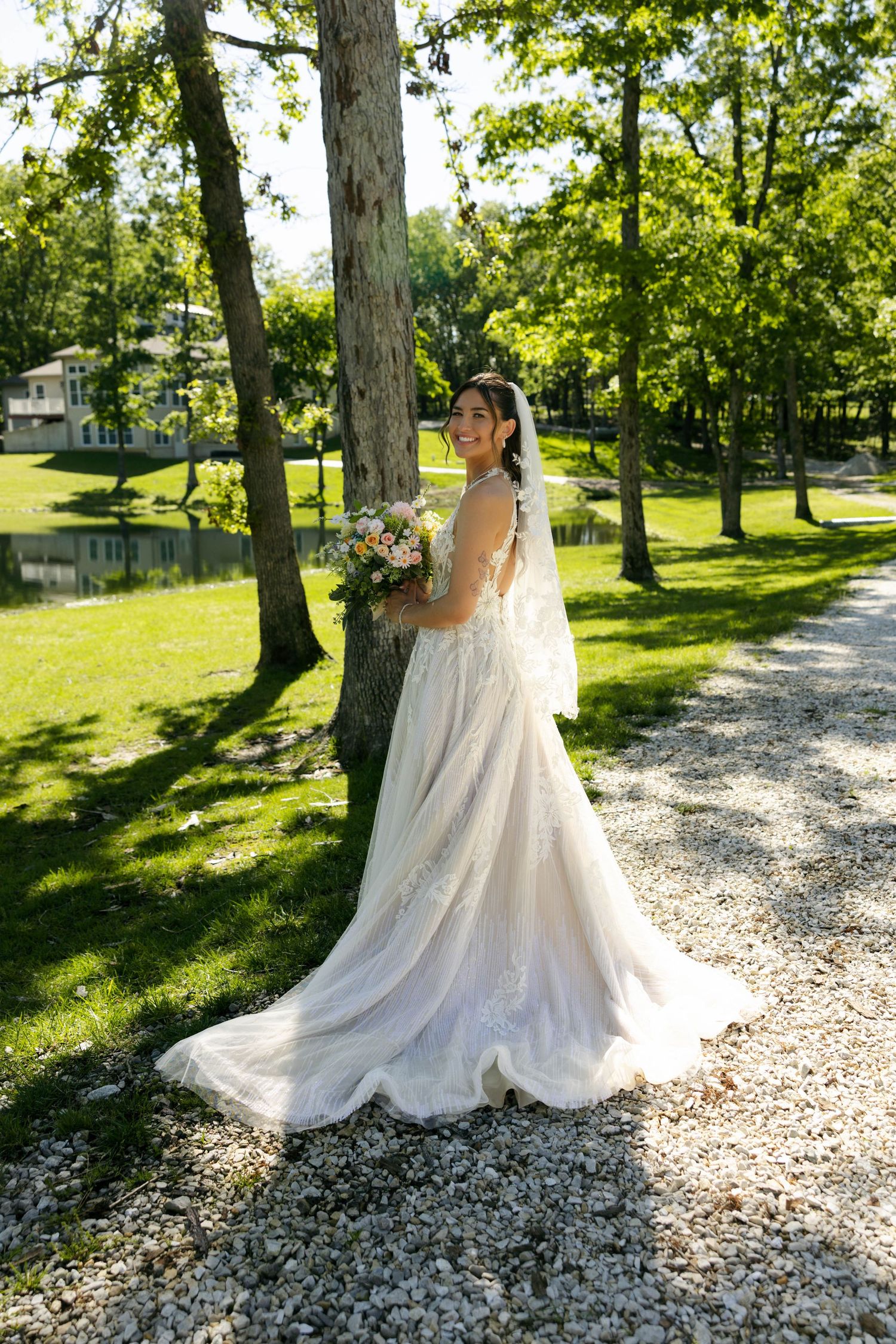 Bride in white gown and veil holding bouquet, smiling in a lush green park with trees and gravel path.