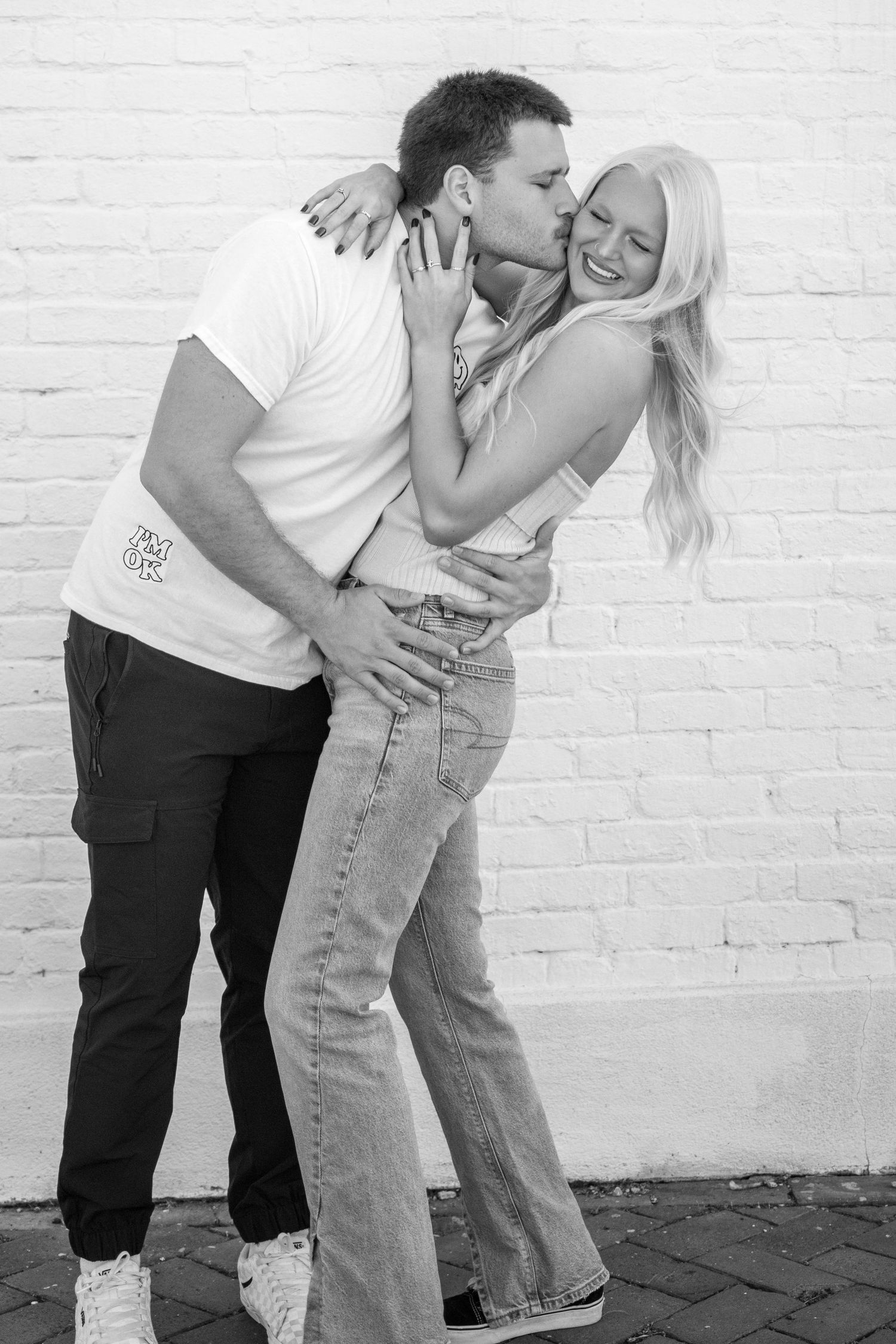 Romantic black and white photo series of a couple embracing against a white brick wall.