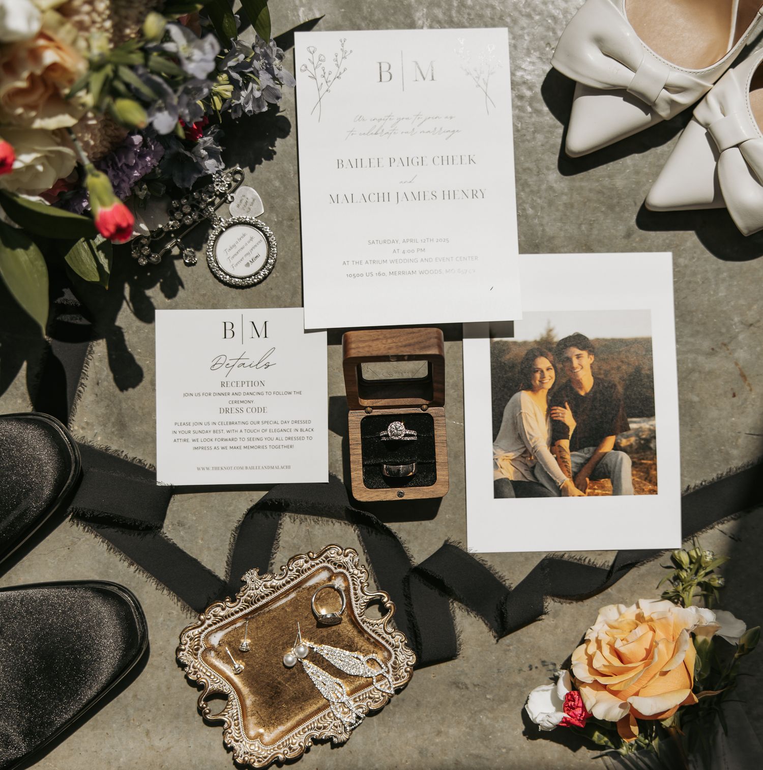 Wedding details flatlay with invitation suite, floral arrangements, vintage frame, and ring box on grey surface.