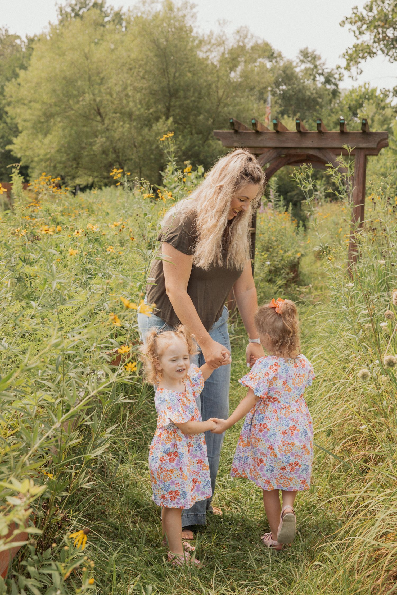 Family walking hand in hand through tall grass and wildflowers near wooden pergola.