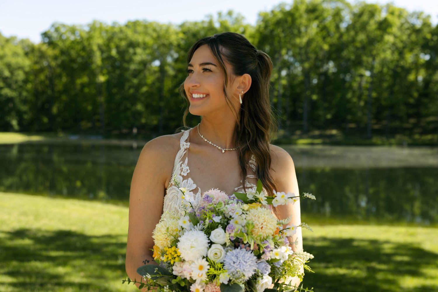 Smiling bride holding white and yellow floral bouquet outdoors near a pond with lush green trees in the background.