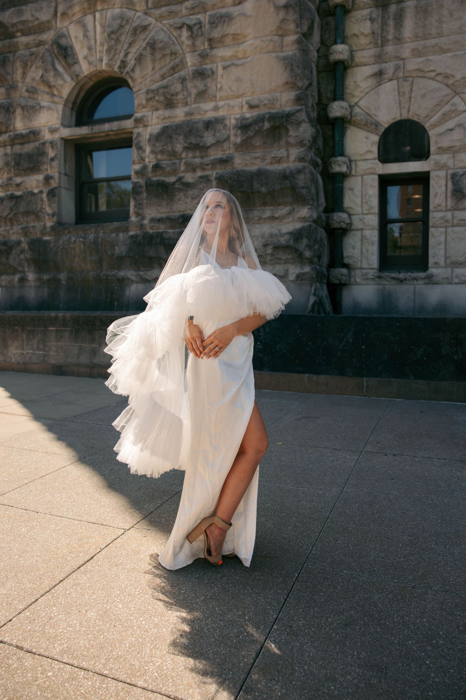 Elegant bride in white gown with tulle sleeves and veil poses in golden sunlight outside stone building.