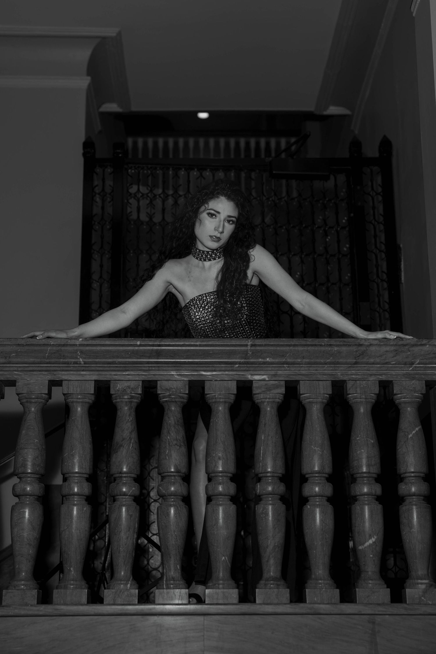 Elegant woman in black dress leaning on ornate balcony railing in black and white photo.