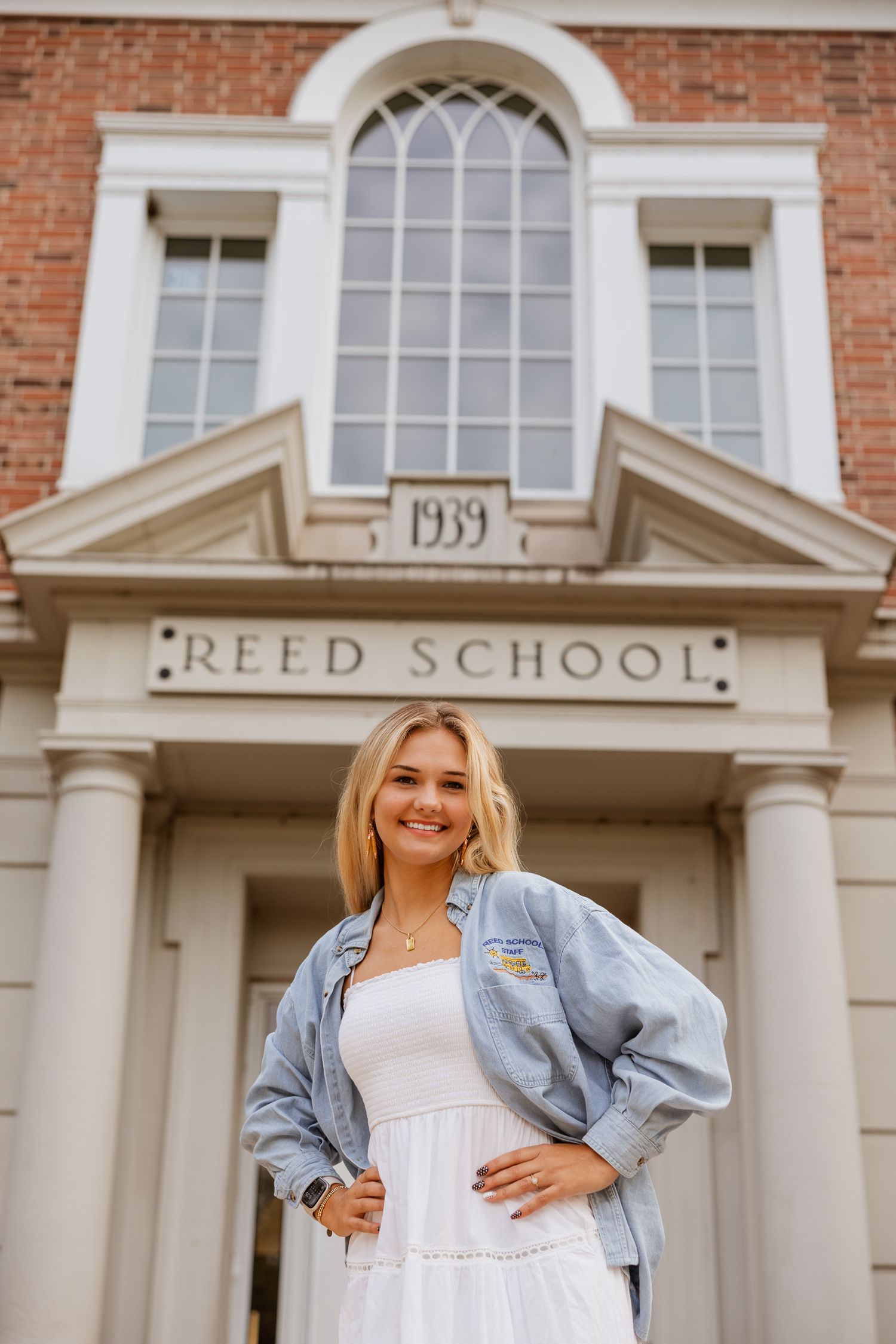 A casual outfit featuring a white dress and denim jacket is showcased in front of a historic school building.