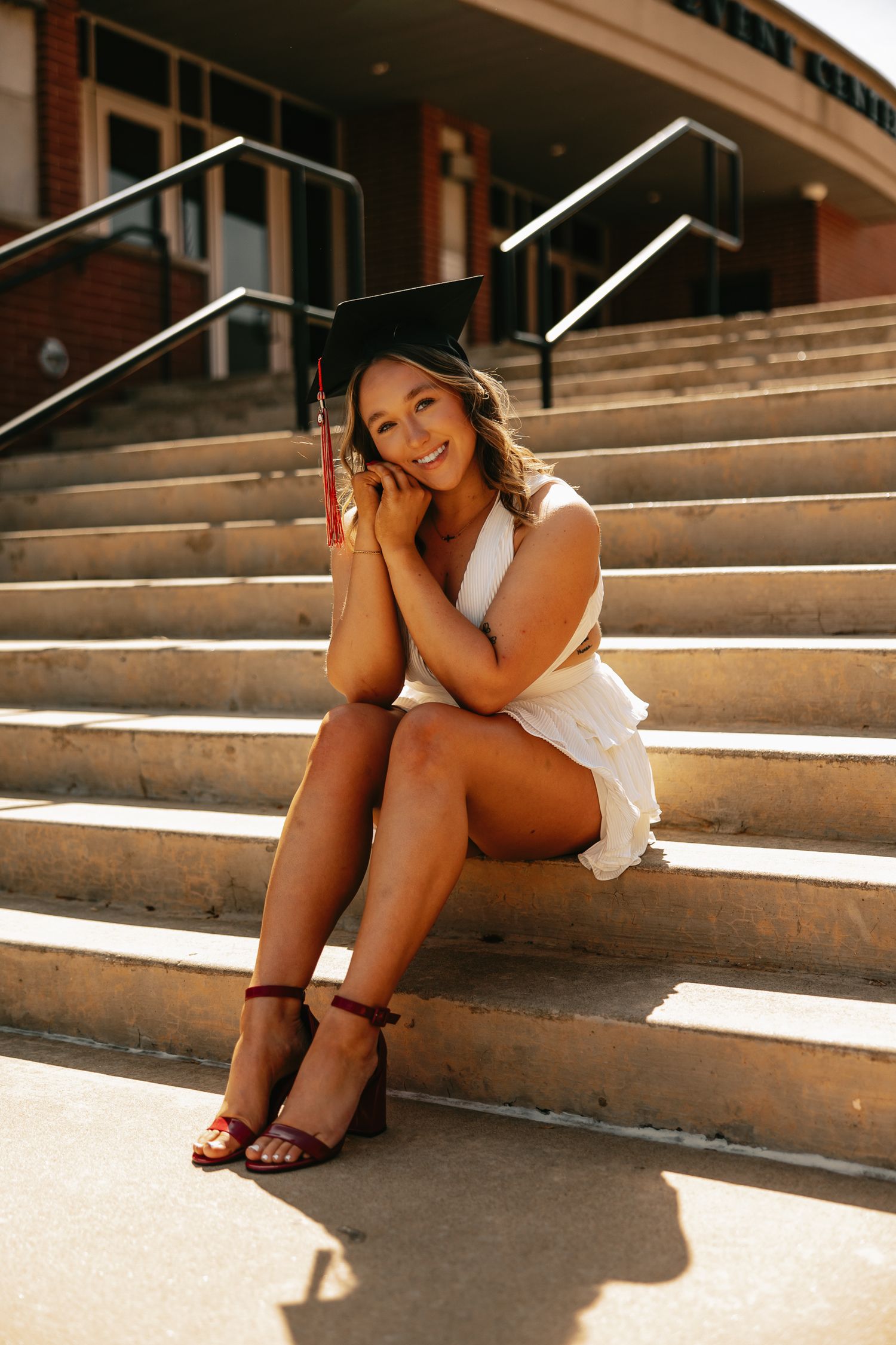 Graduate in white dress lounges on concrete steps in sunlit outdoor setting.