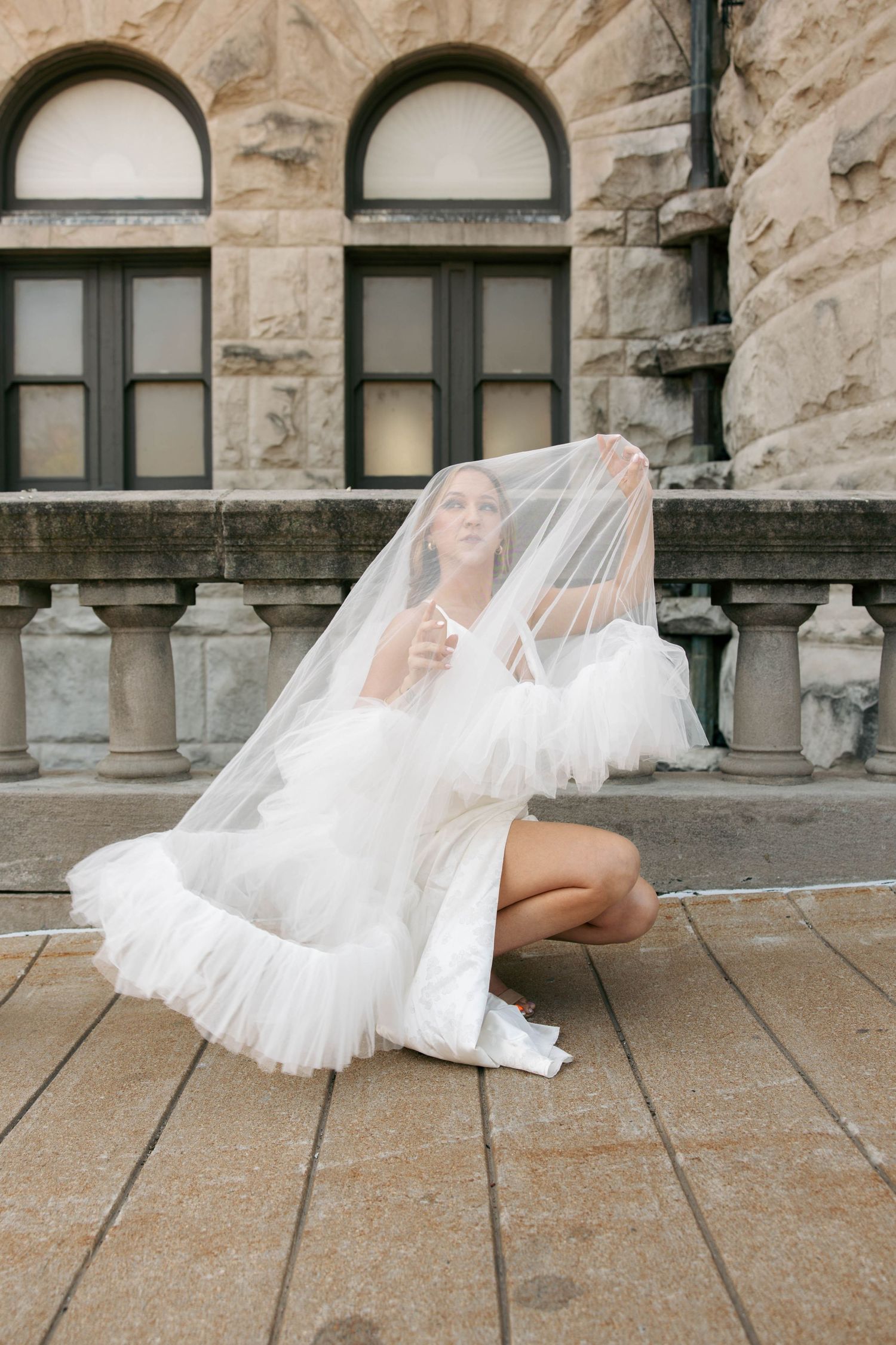 Bride in white tulle mini dress sits on stone steps, lifting veil dramatically before a classical stone building.