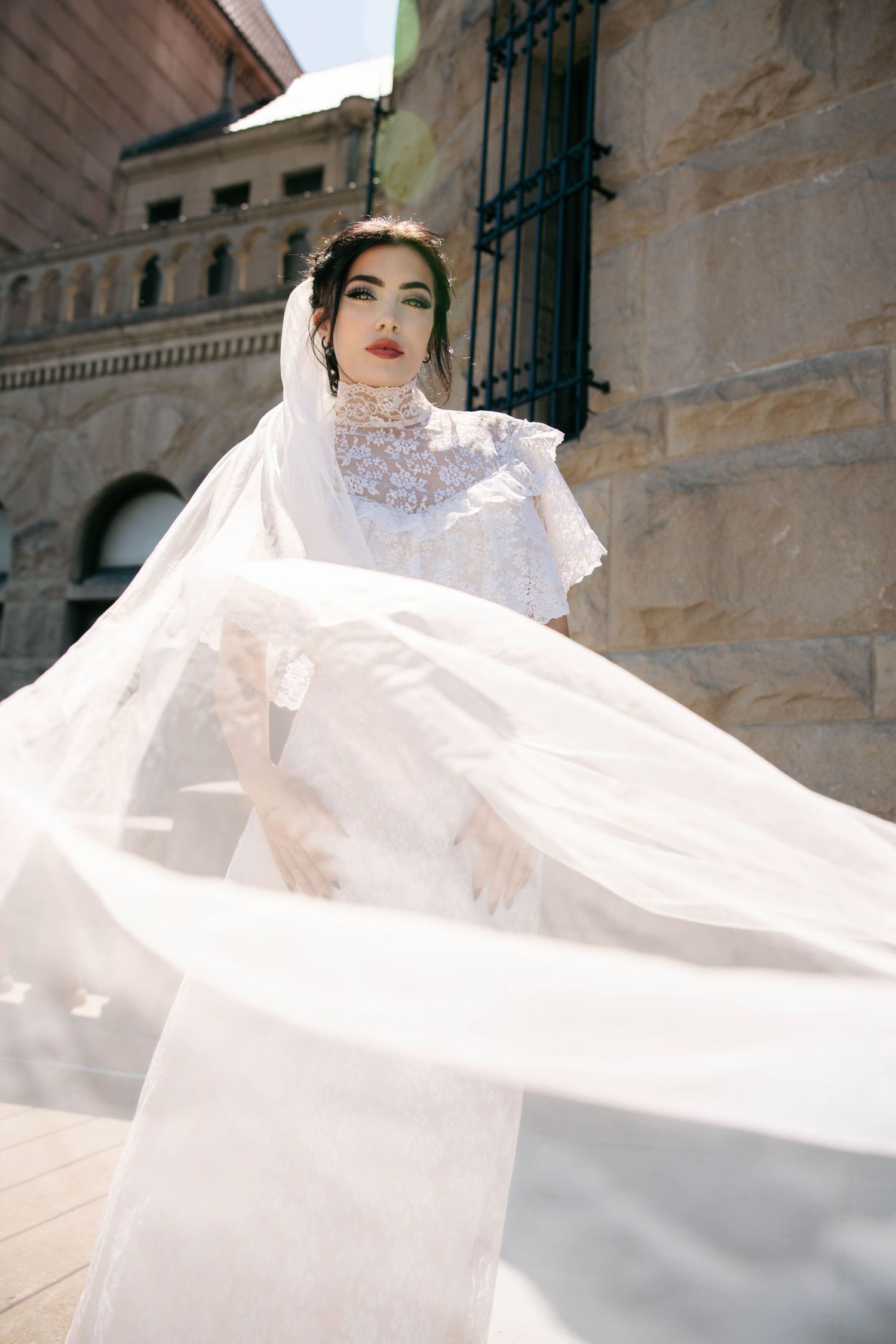 Bride in high-neck lace wedding dress with billowing veil flowing in the wind outdoors.