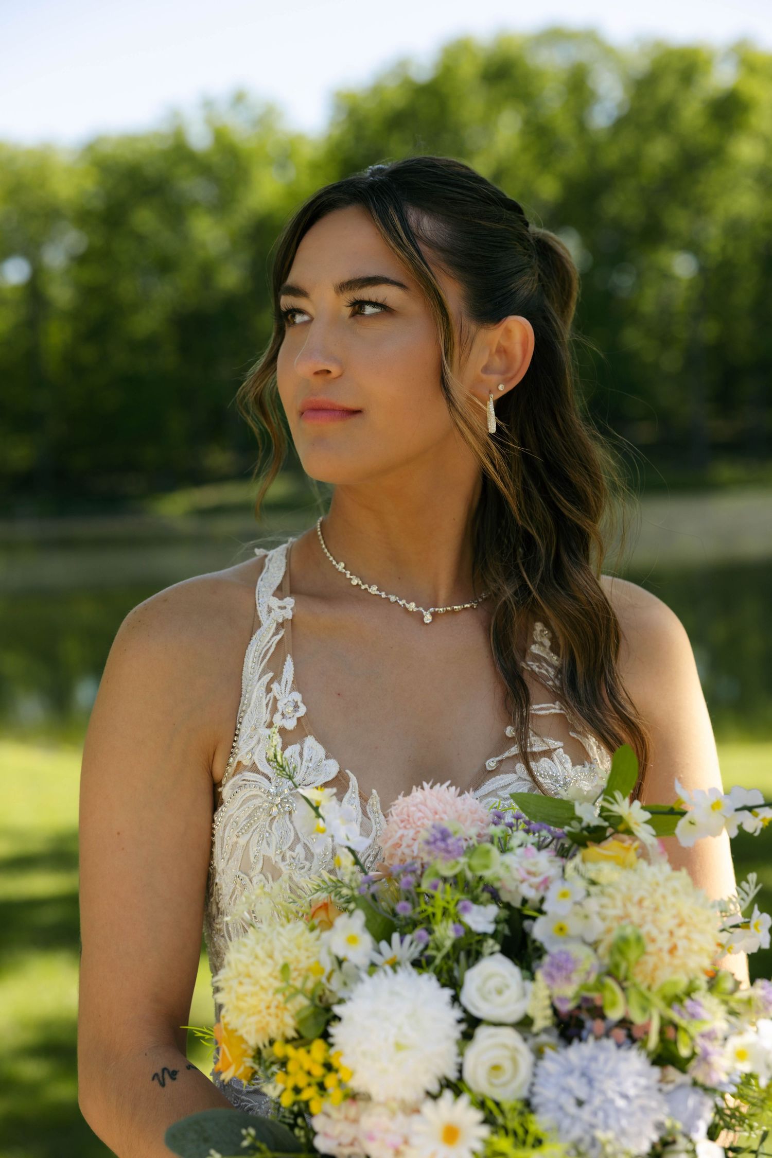 Bride in lace wedding dress holding colorful wildflower bouquet outdoors near water, wearing pearl necklace and earrings.