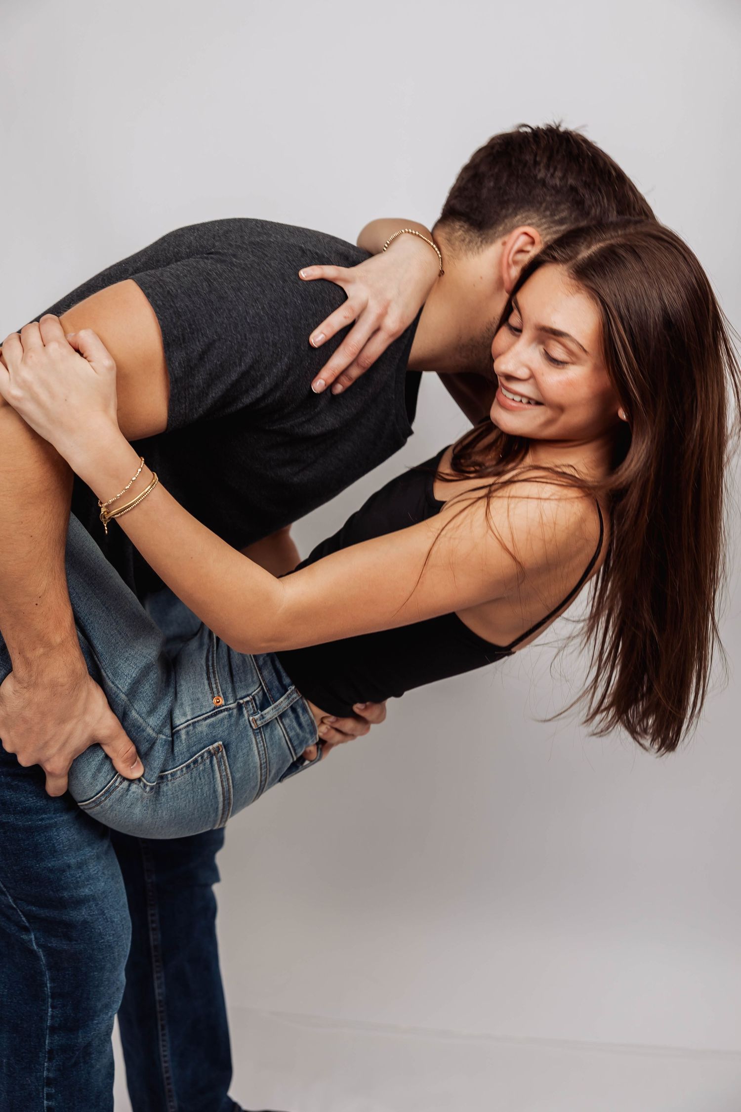 Playful studio pose of couple in jeans and dark tops against white background.