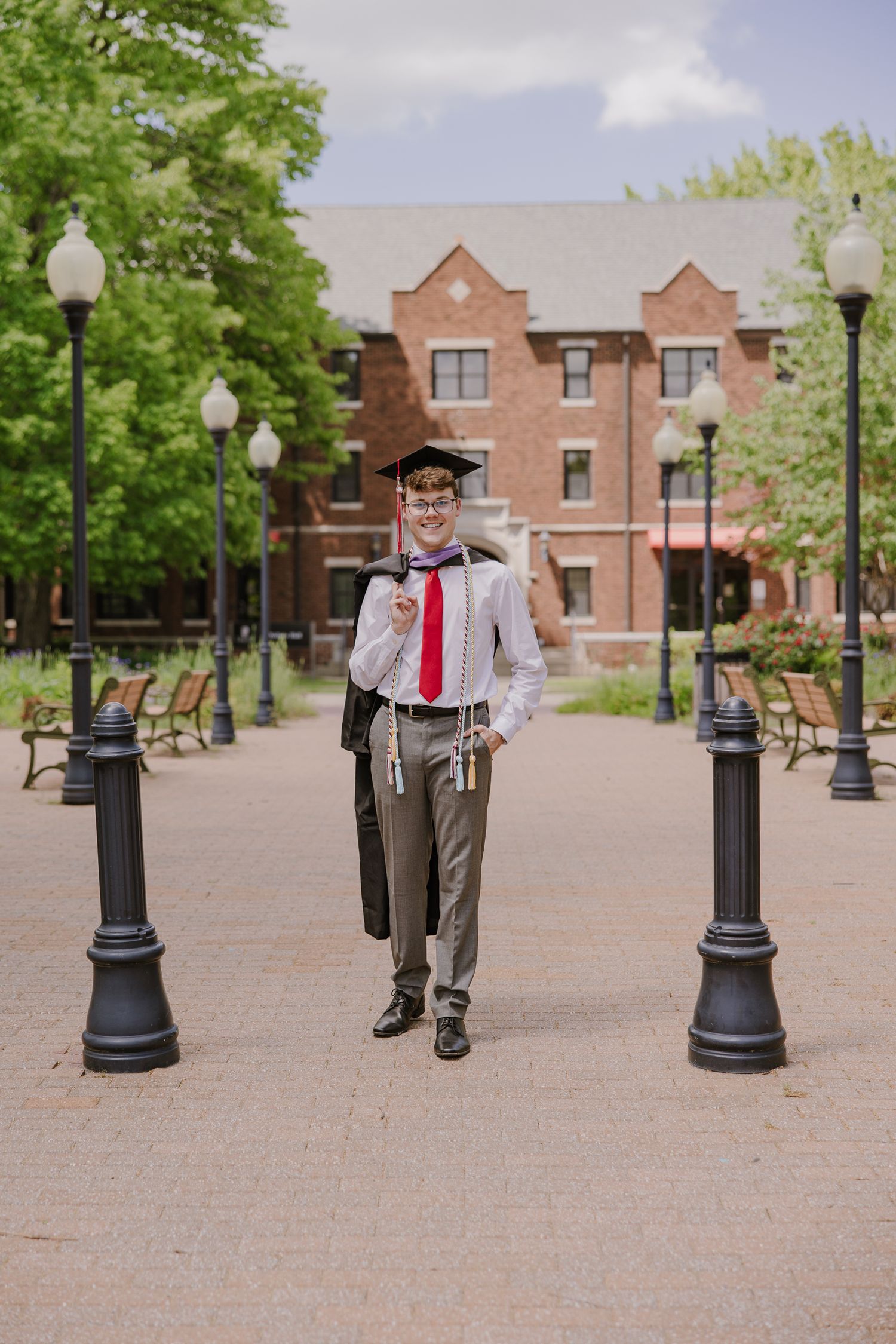 A graduate in academic regalia poses playfully between lamp posts on a brick plaza.