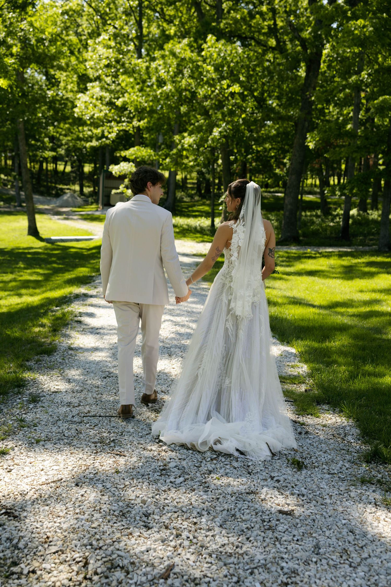 A bride and groom walk hand in hand down a gravel path surrounded by lush green trees on their wedding day.