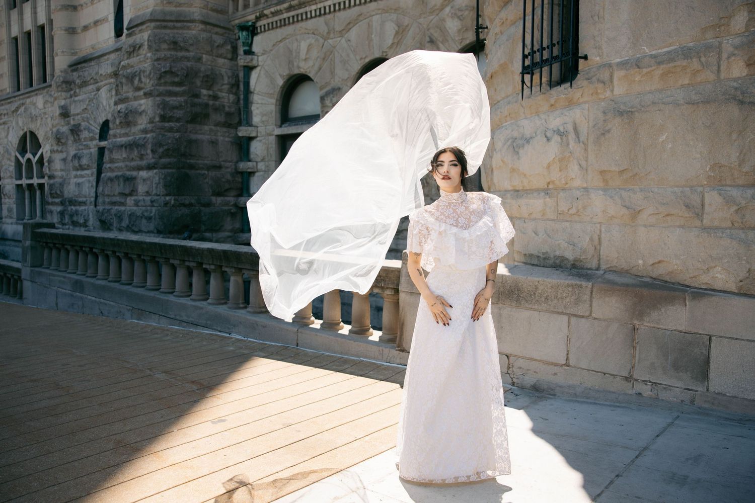 Bride in elegant white gown with billowing veil caught in wind stands before a historic stone building.