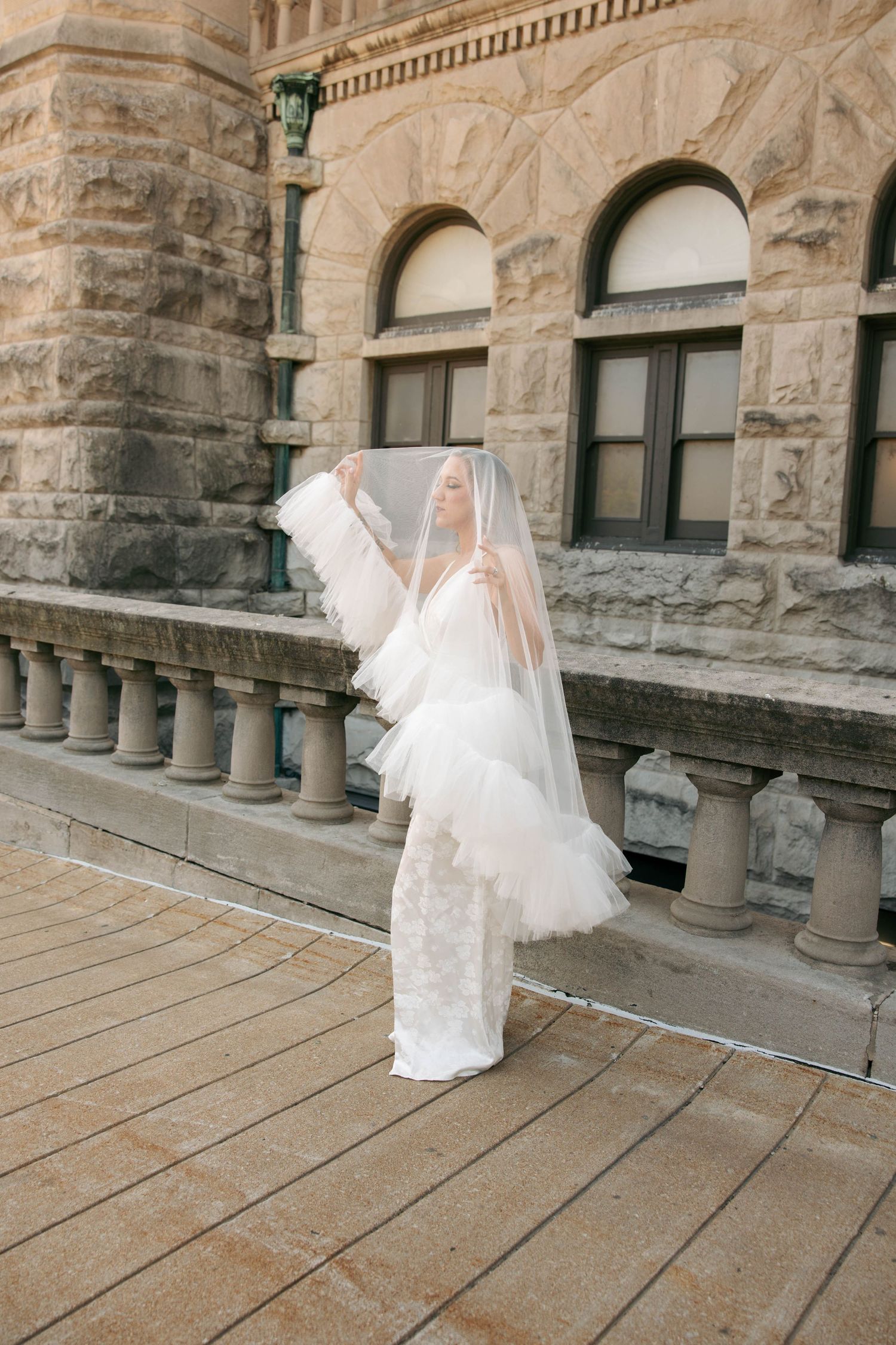 Bride in white lace gown and veil poses gracefully on stone balcony of historic building.