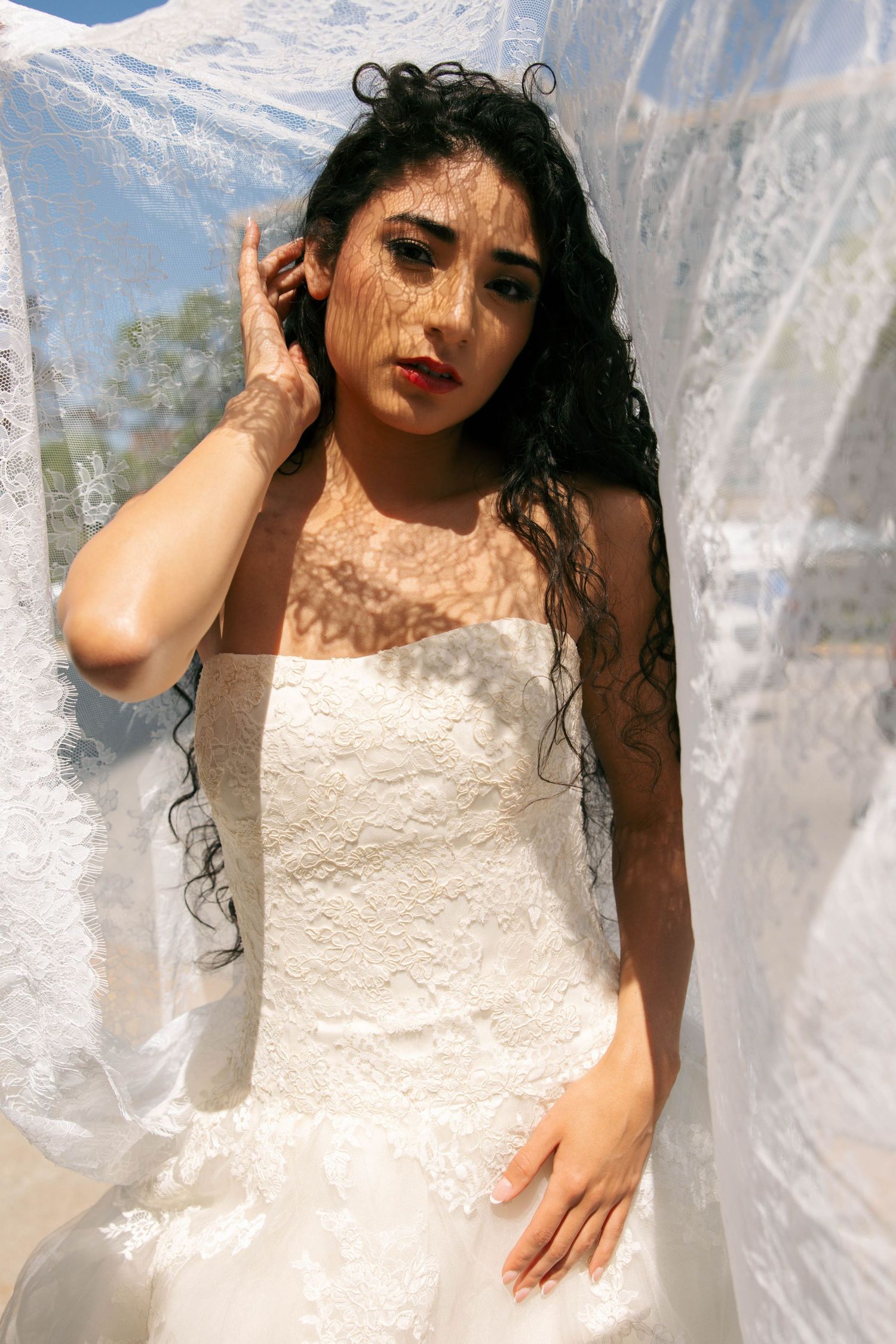 Beautiful bride in strapless lace wedding gown posing behind sheer fabric with dappled sunlight on face.