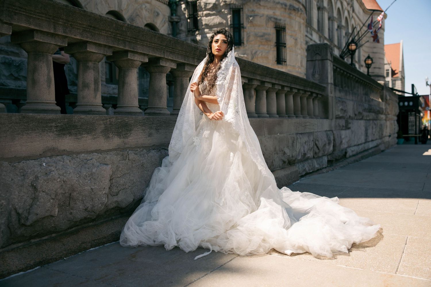 Elegant bride in a flowing white tulle wedding gown with cape train posing on a historic city street.