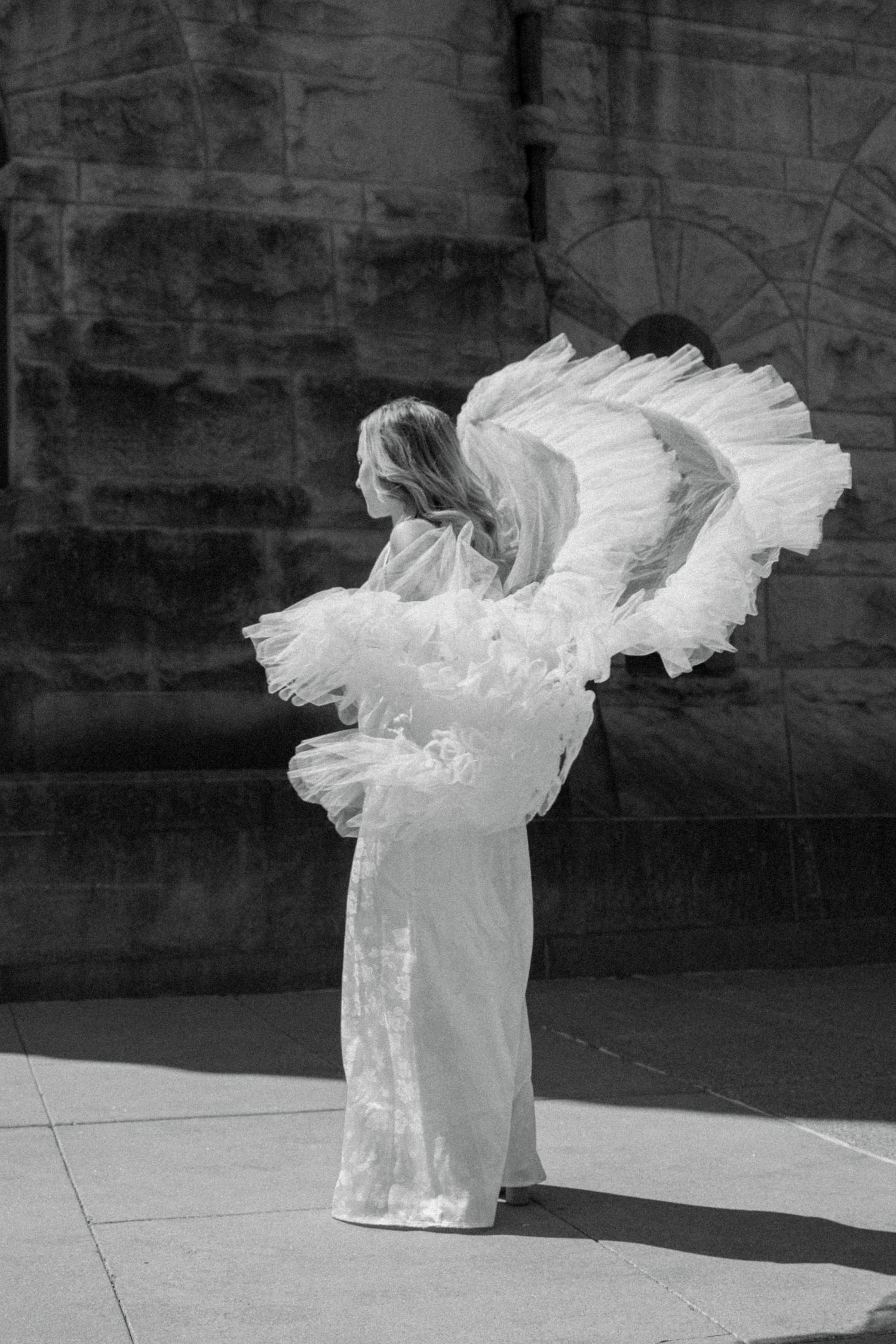 Black and white photo of a woman in white dress with large angel wings posing outdoors near a stone building.