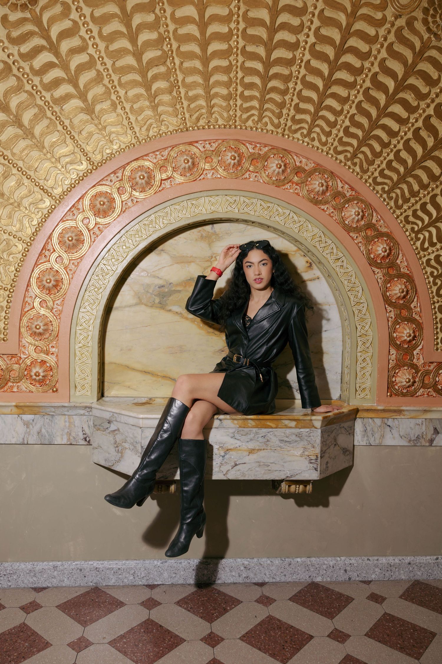 Woman in black leather outfit and knee-high boots poses in ornate golden arched alcove with decorative ceiling.
