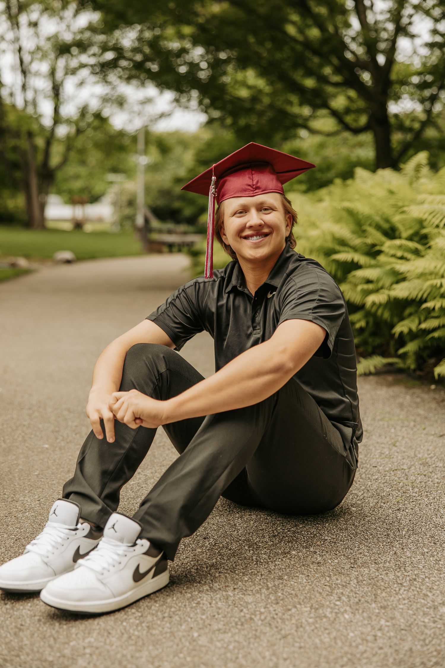 Graduate in black outfit and maroon cap relaxes on outdoor path.