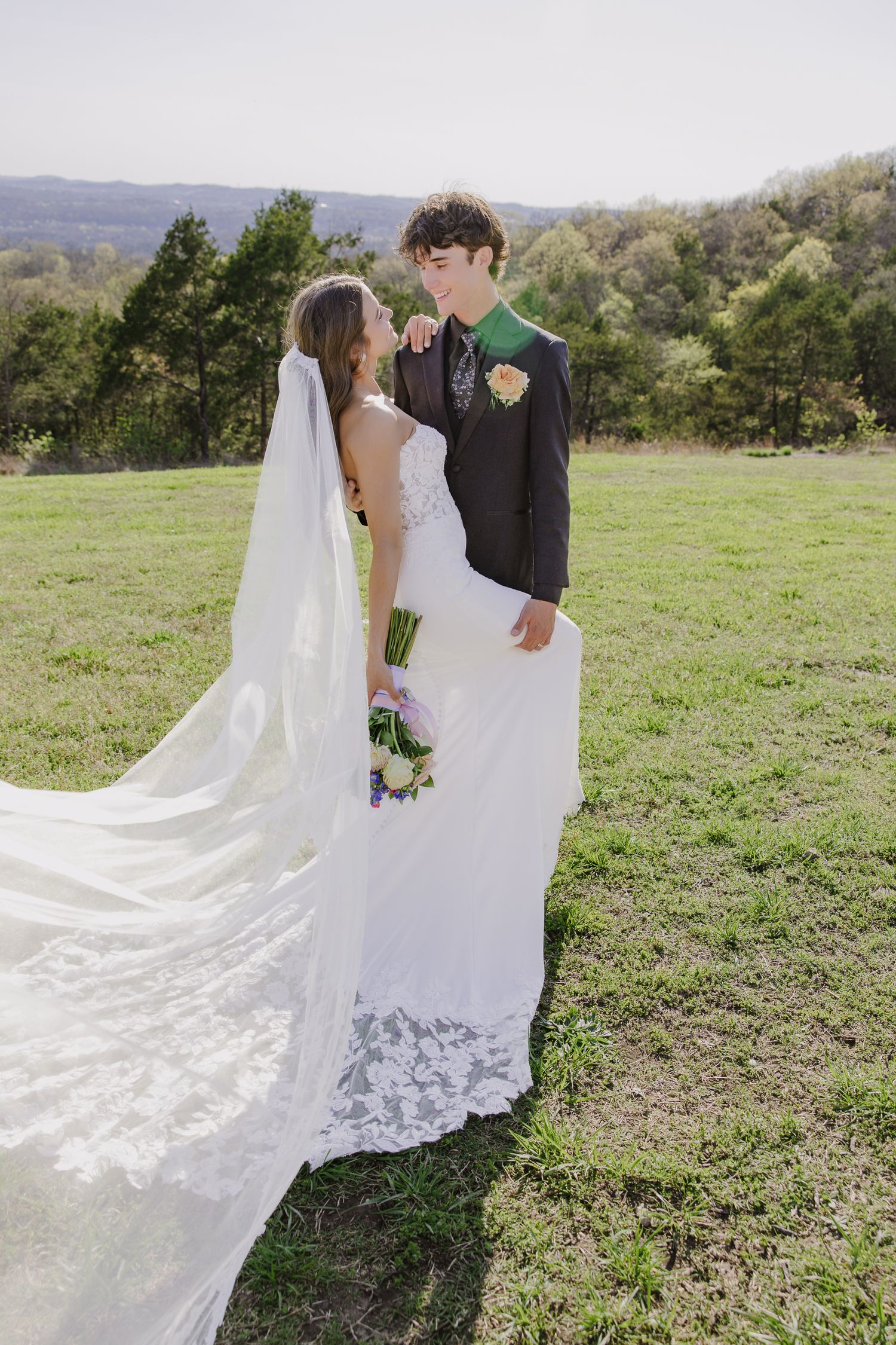 Romantic wedding portrait on a hilltop with flowing lace veil and spring greenery.