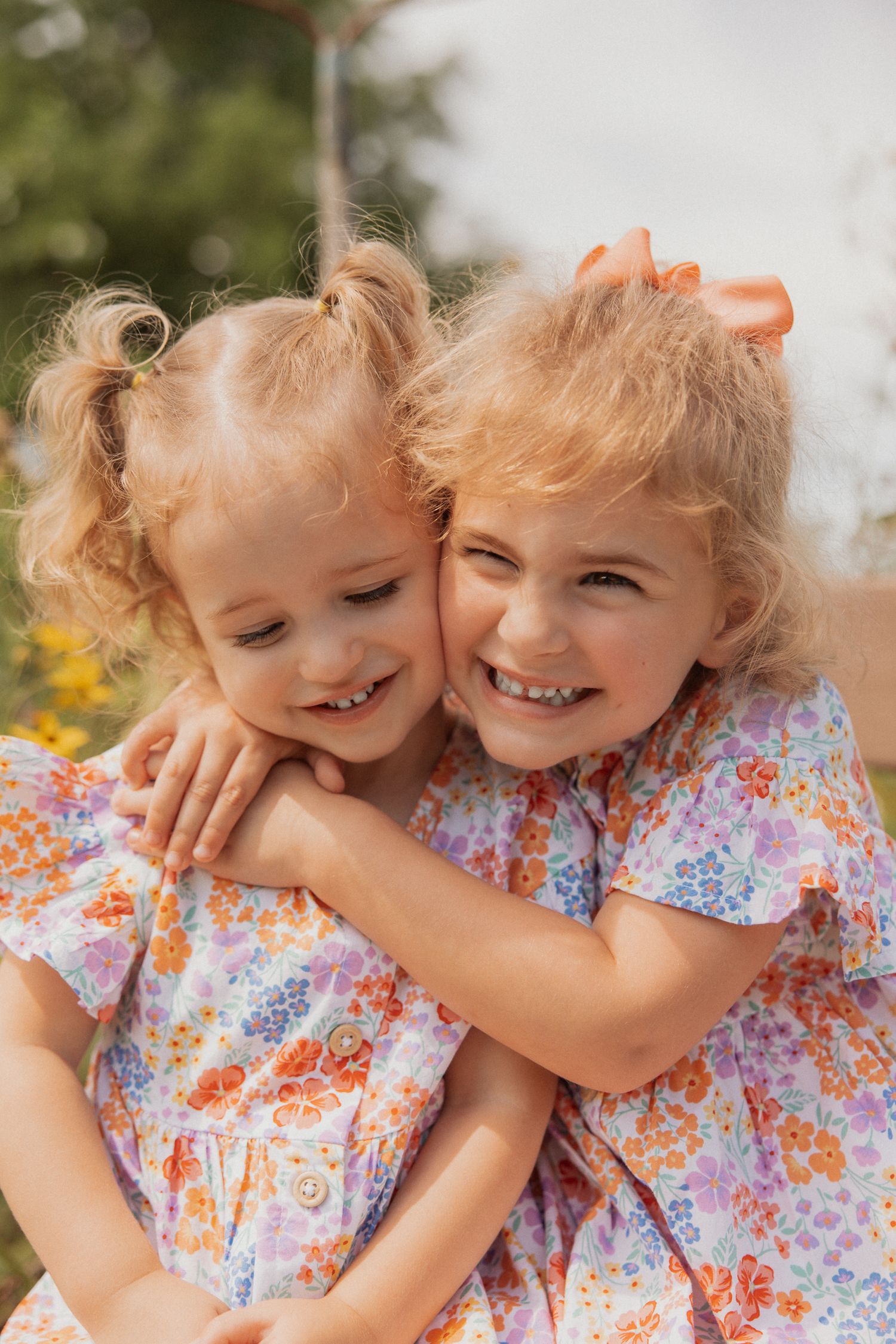 Two children in matching floral dresses share an affectionate moment outdoors.