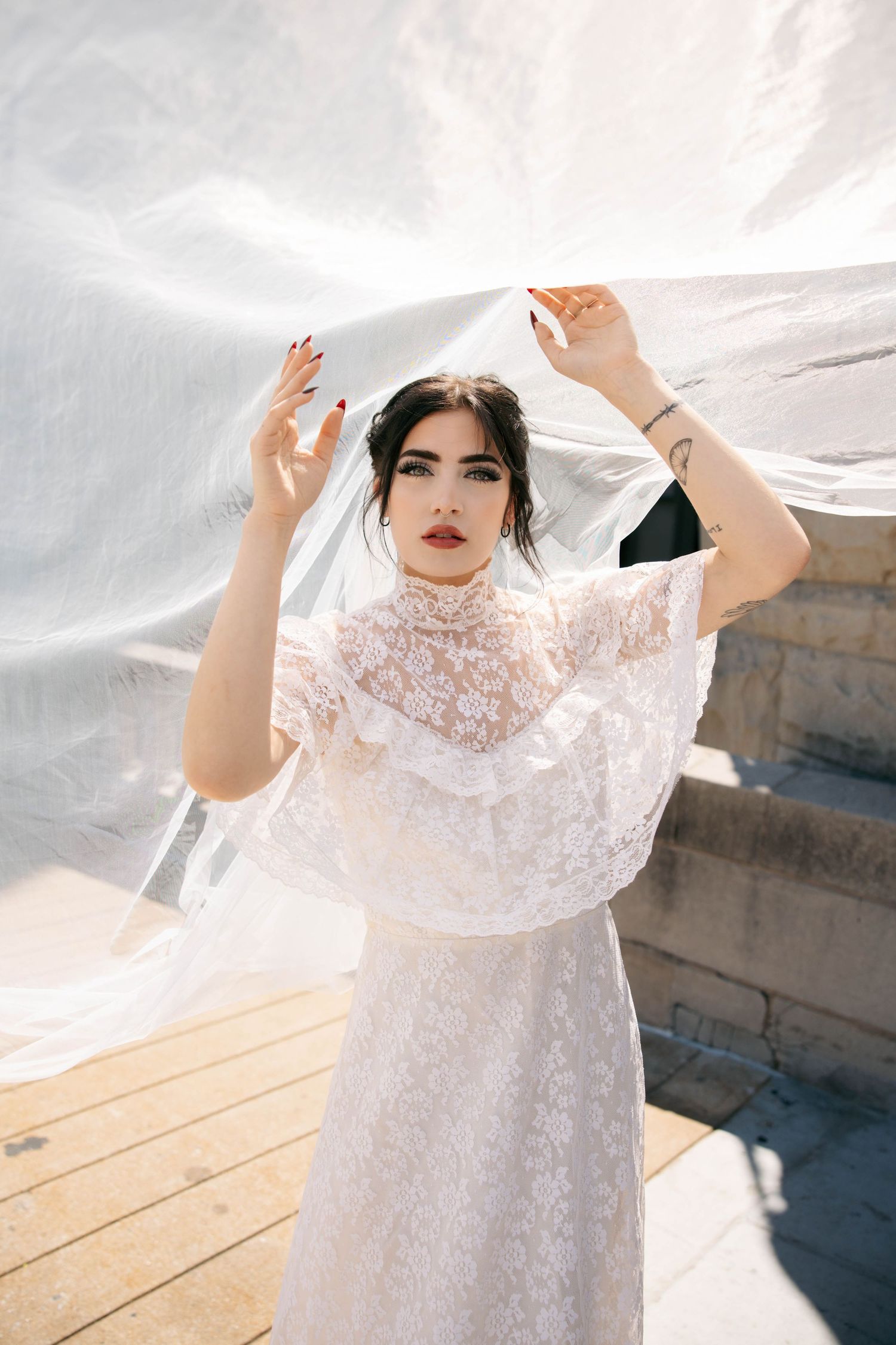 Bride in white lace high-neck dress holds flowing veil overhead outdoors in sunlight.