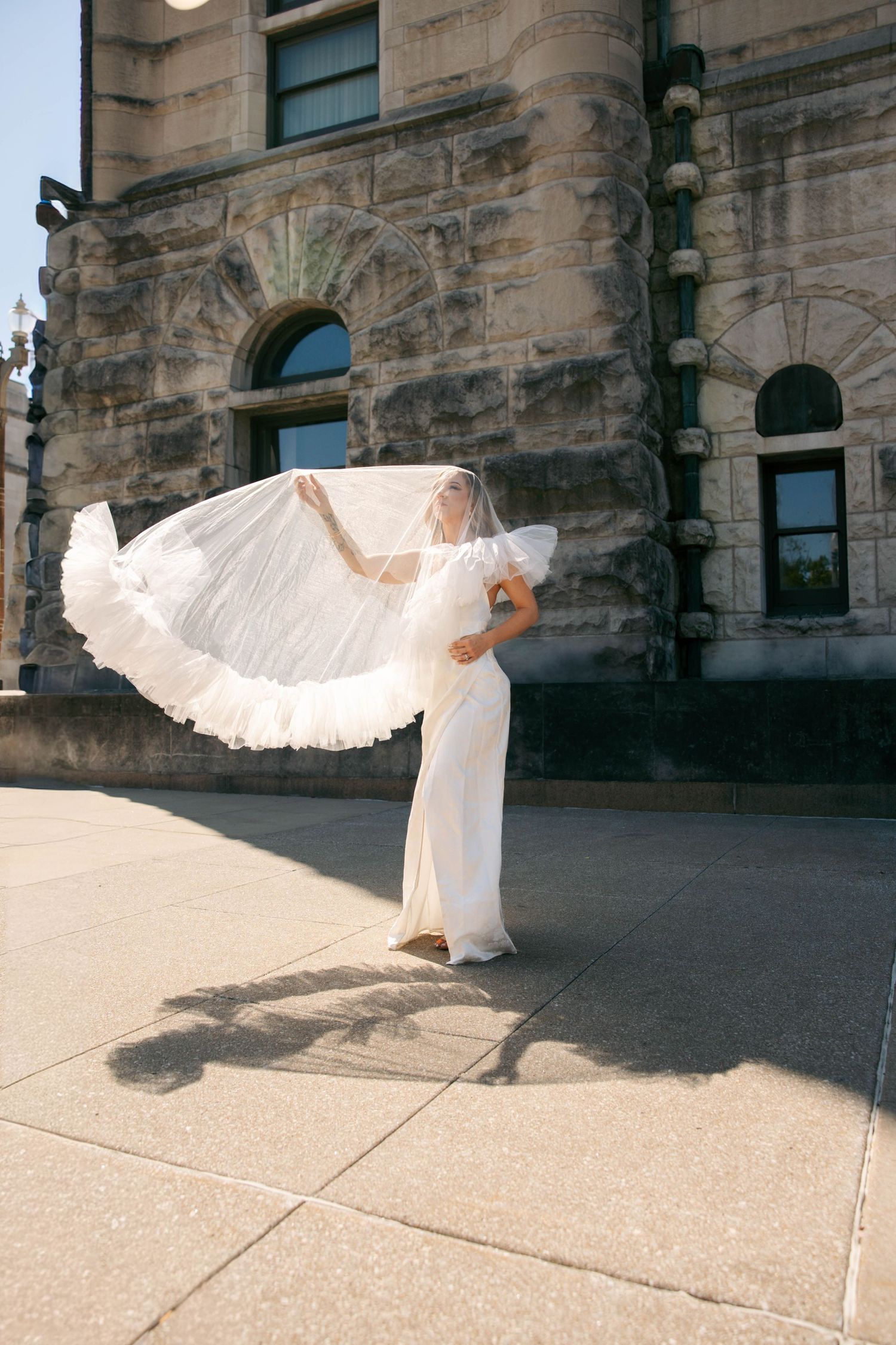 Bride in white gown tosses flowing veil outdoors near stone building on sunny day.