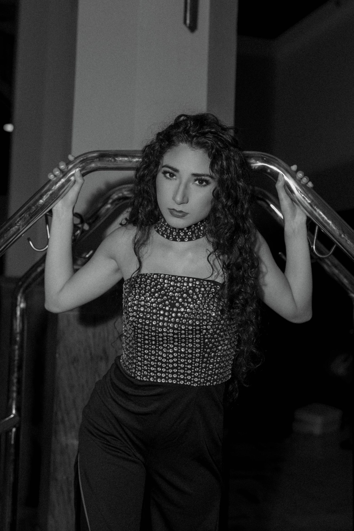 Black and white photo of a young woman with curly hair posing on stairs, wearing a sequin top and choker necklace.