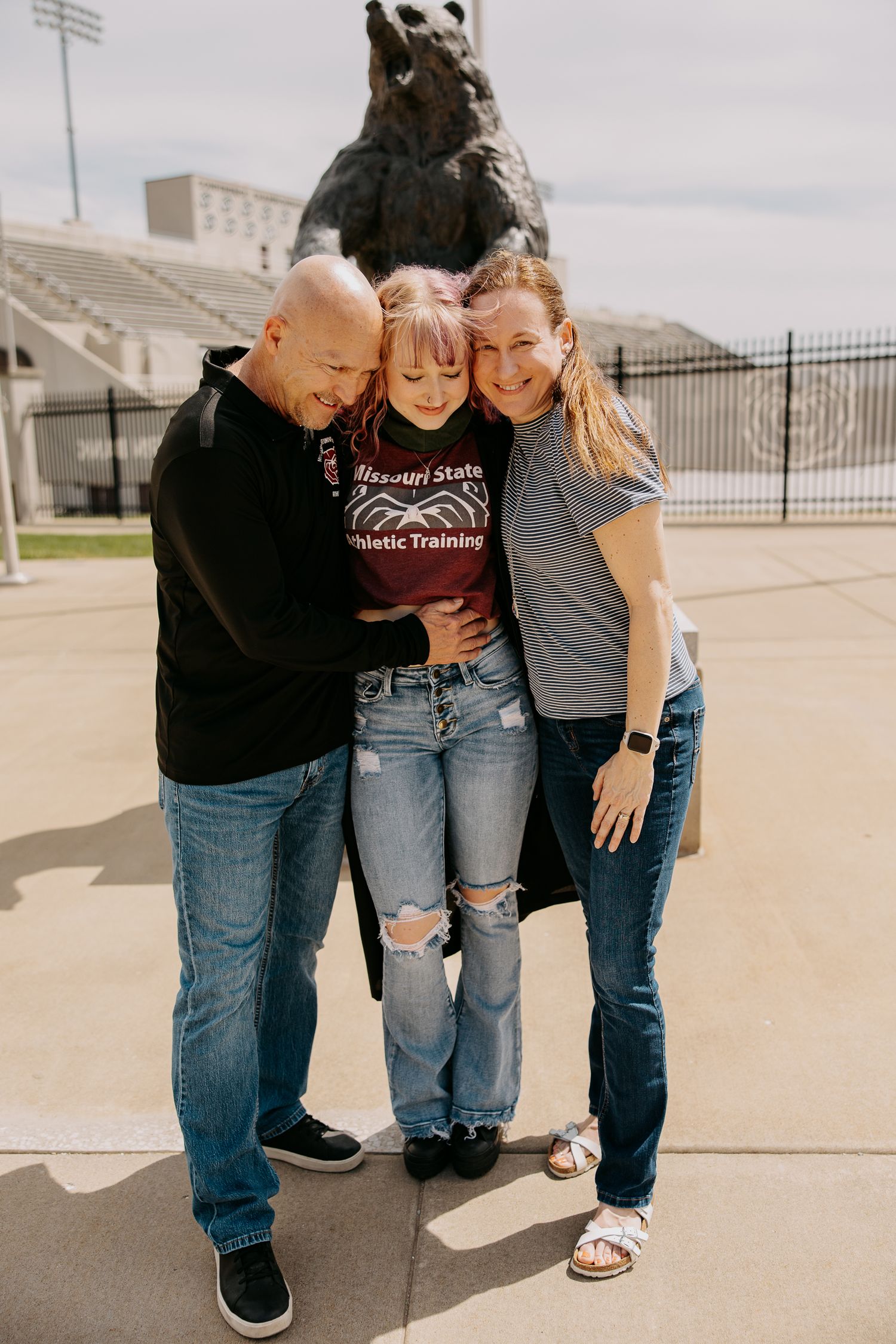 Three people in casual wear share a close moment on a concrete surface.