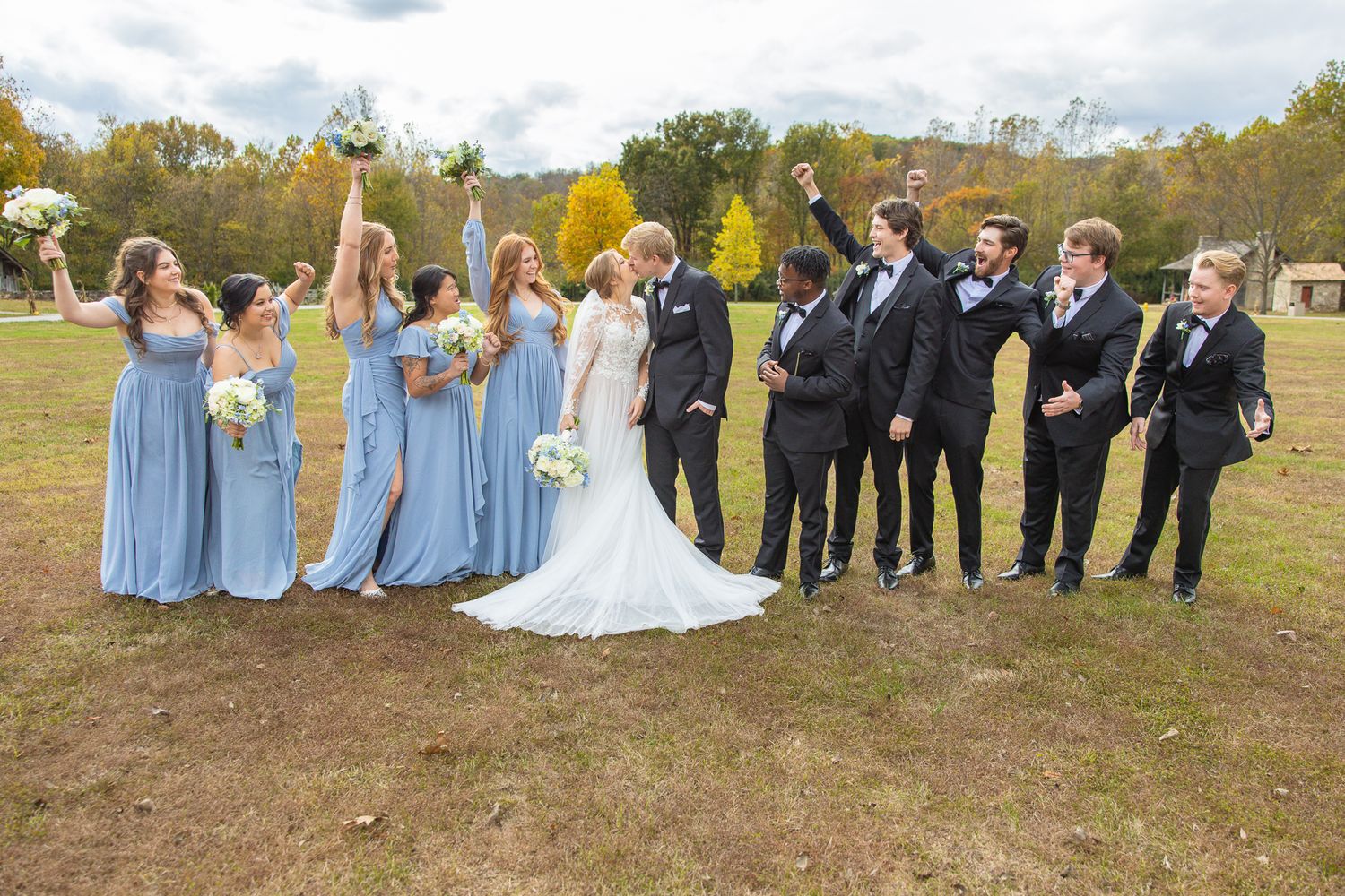 Wedding party in light blue dresses and dark suits poses outdoors on autumn day.