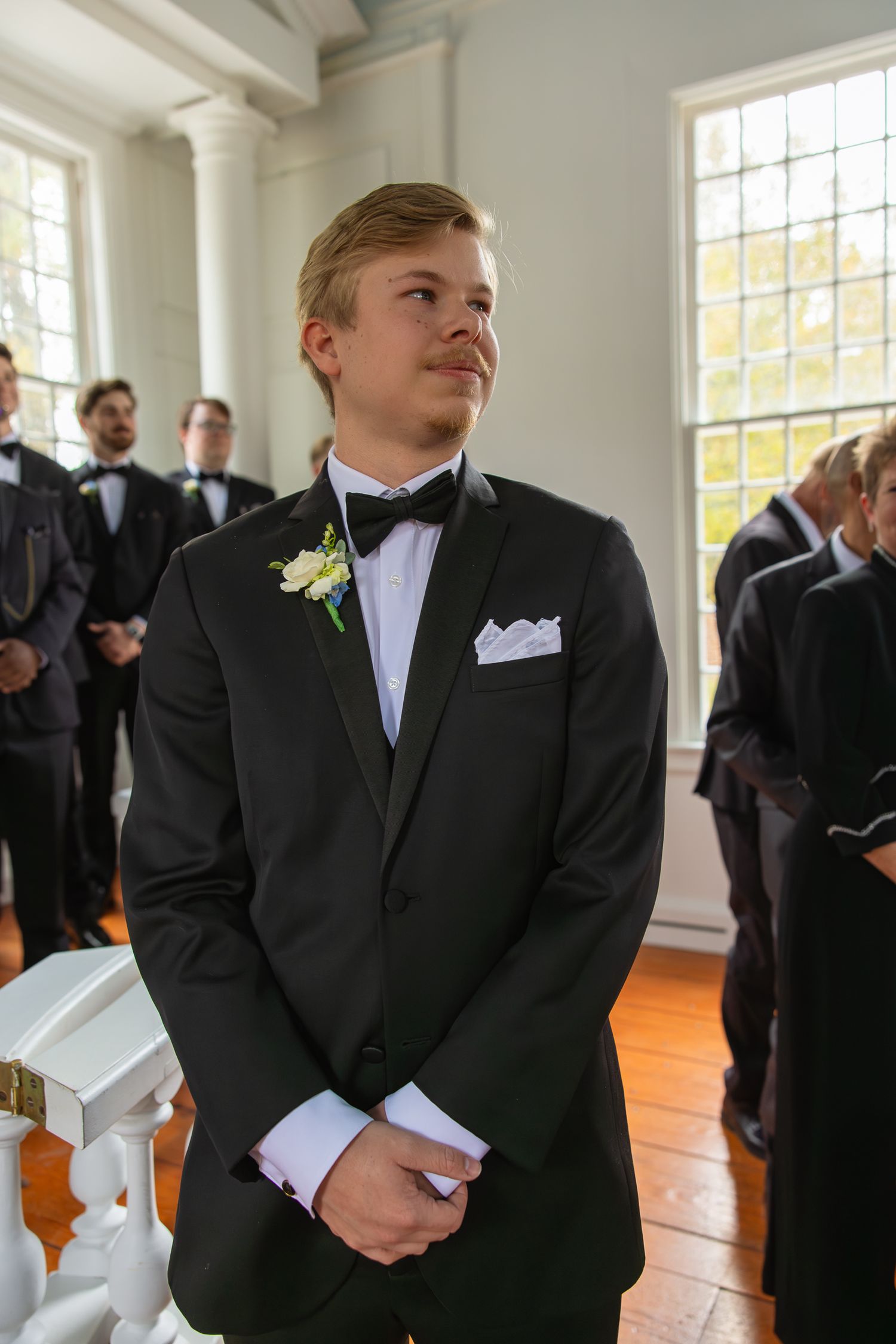 Groomsmen in black suits standing in an elegant white room with large windows.