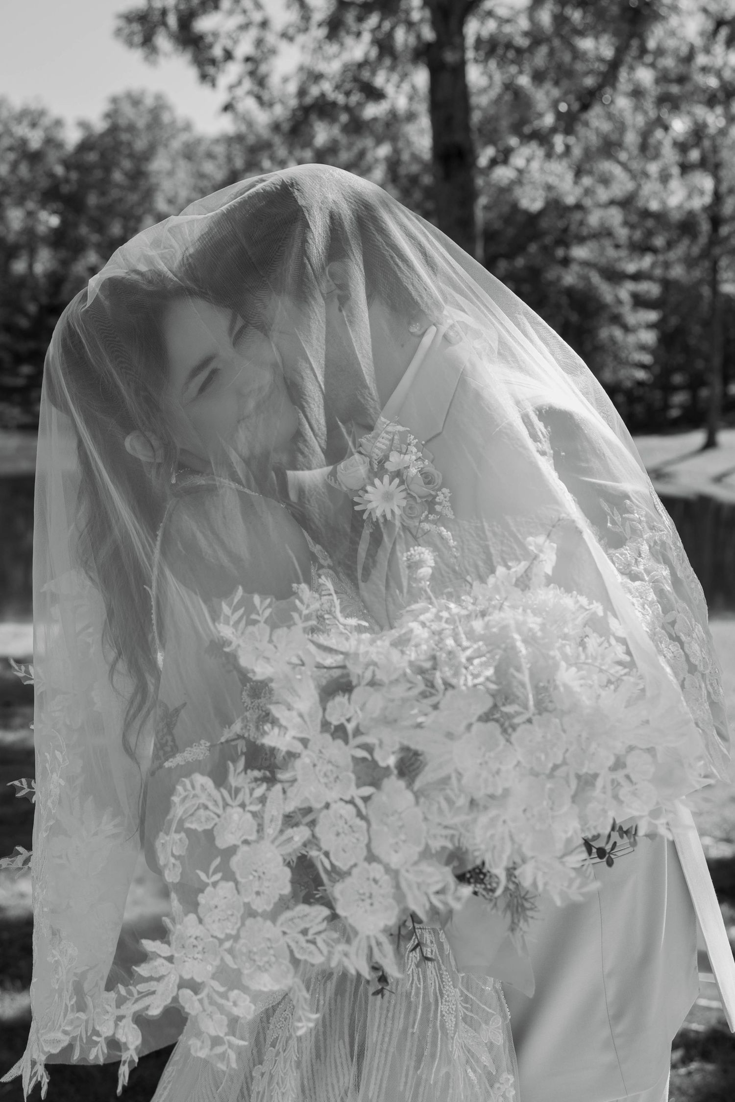 Black and white bridal portrait of a bride under a lace veil holding a floral bouquet outdoors.