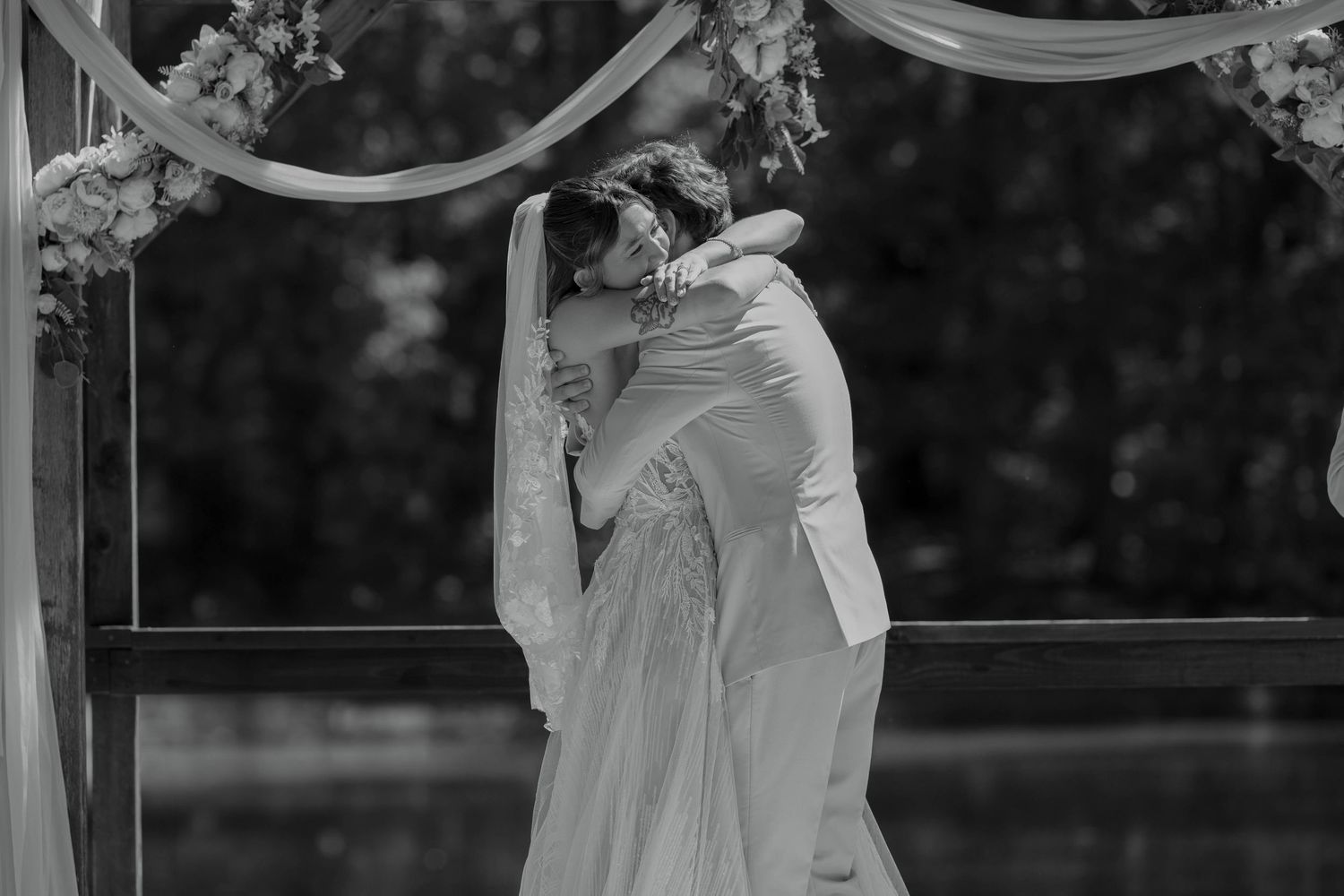 Black and white photo of a bride and groom sharing a romantic embrace under a floral arch at their wedding ceremony.