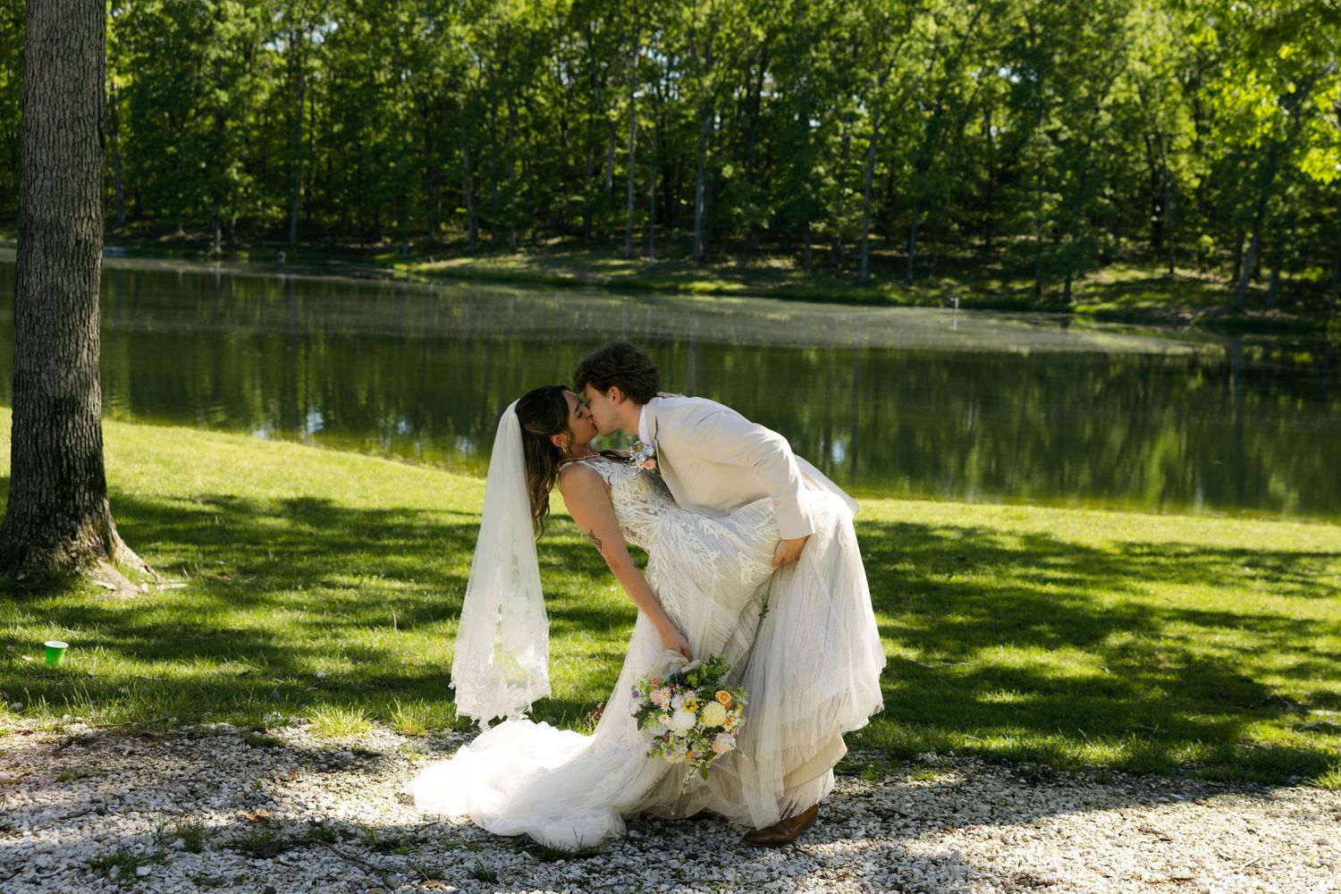 Bride and groom sharing a romantic kiss near a serene pond surrounded by lush green trees on their wedding day.