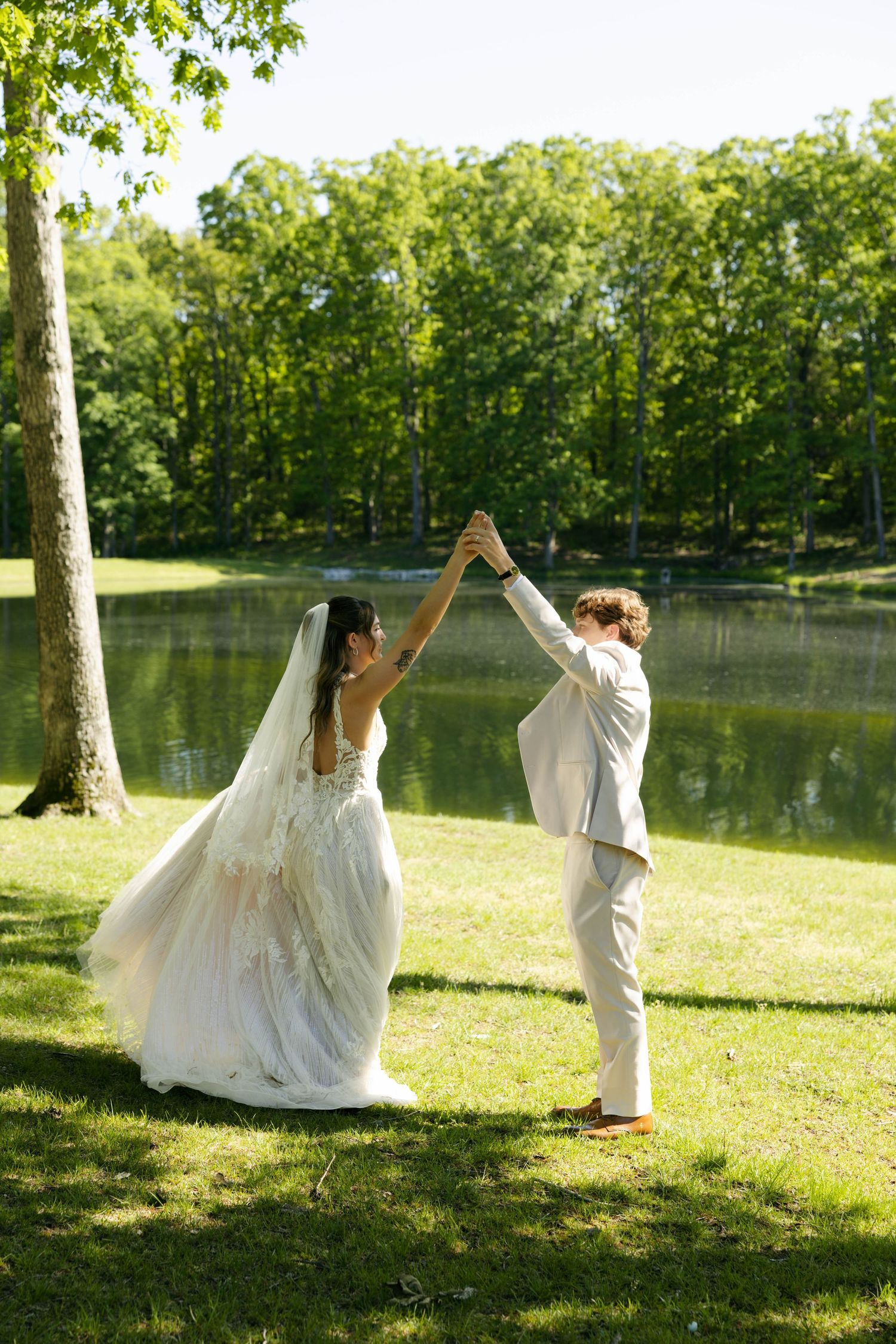 Bride and groom dancing on green lawn by a serene lake, surrounded by lush trees on a sunny wedding day.